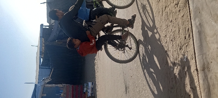 A man stands with a child on a bicycle. The man is dressed in a black top, while the child wears a red jacket. They are positioned on a paved area alongside a sandy patch marked with footprints. In the background, there is a structure covered with dark material. The sunlight casts shadows of the people and the bicycle on the sandy ground.