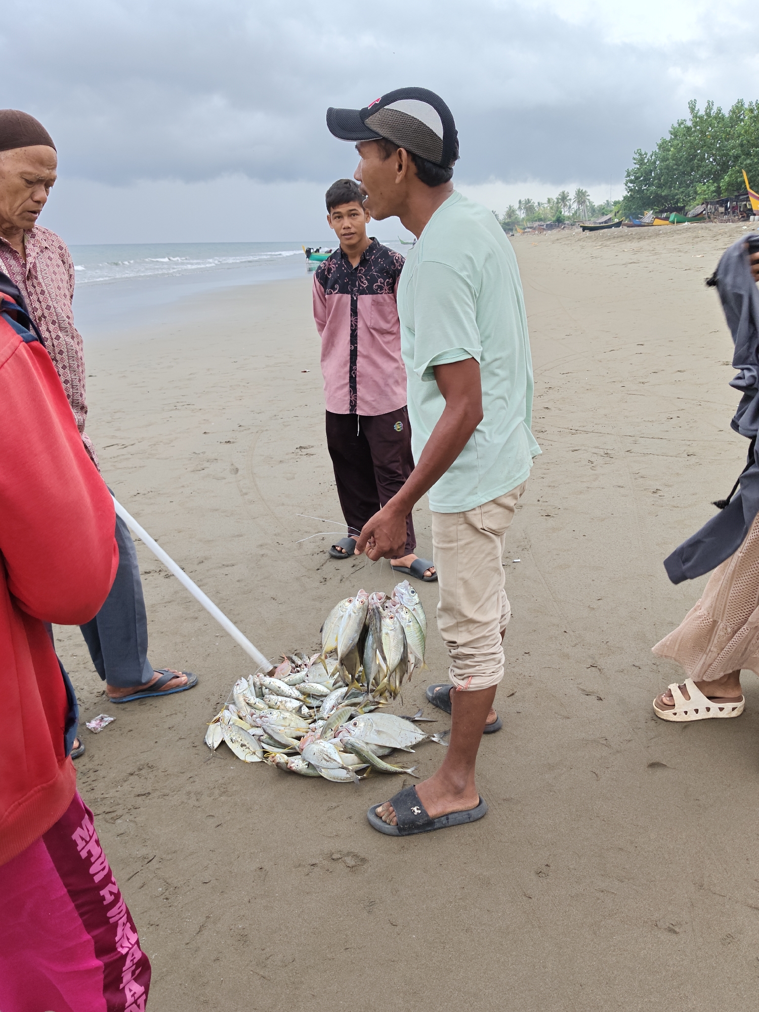 A group of people are gathered on a sandy beach in Curehtunong, Indonesia. The sky is overcast. In the foreground, a pile of freshly caught fish is displayed on the sand. A man wearing a light blue t-shirt and beige pants stands next to the fish, holding a string of smaller fish. To his left, a younger person in a pink and black patterned shirt and dark pants stands looking out towards the ocean. Further to the left, an older man wearing a brown skullcap and a patterned shirt is also looking towards the ocean. On the right side of the image, the lower half of another person's body is visible, wearing a long, beige, lacy skirt and white sandals. The ocean is visible in the background with a few small boats on the water. Trees and a few simple structures are visible along the coastline in the distance.