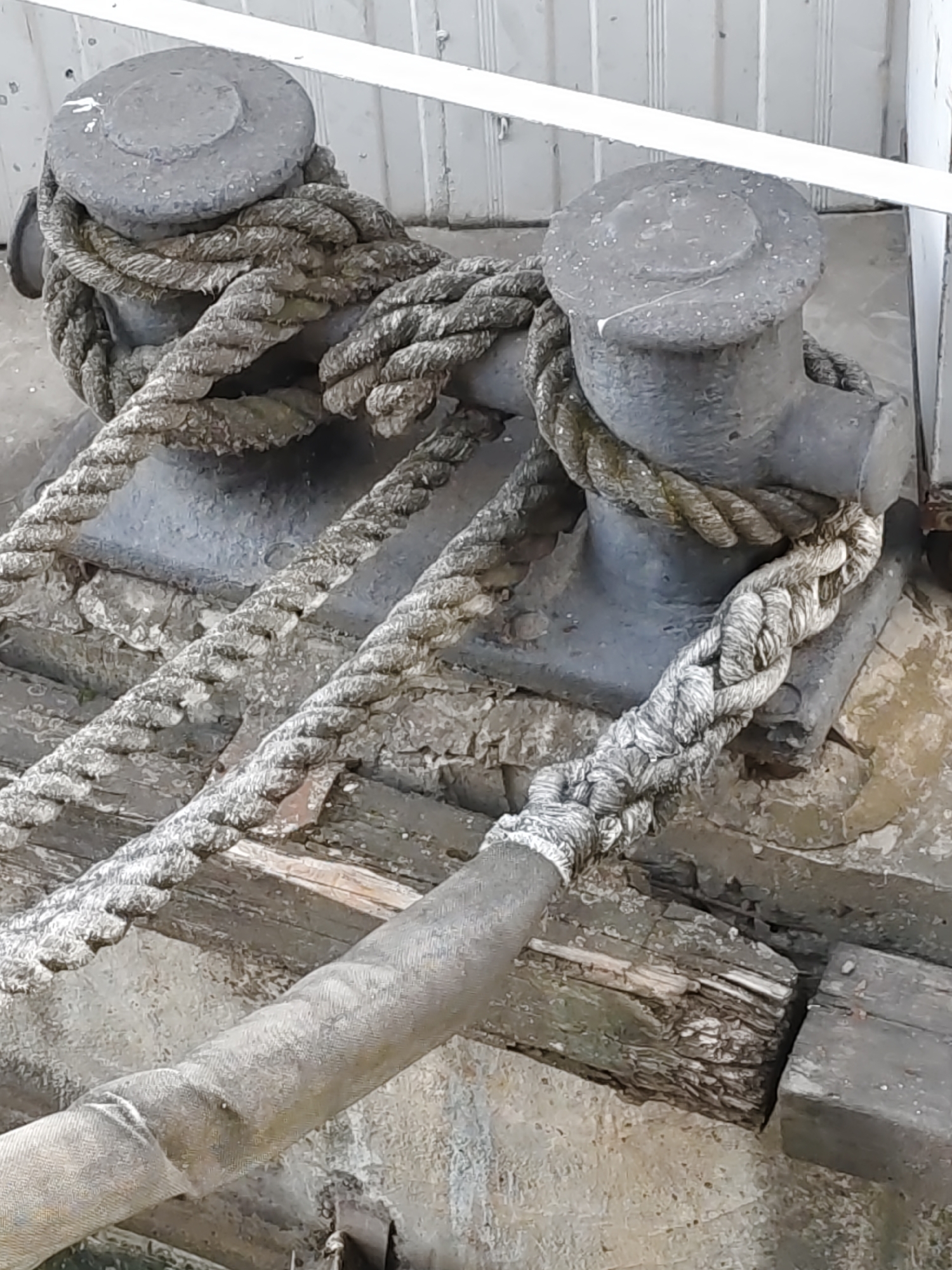 The image shows two metal bollards on a dock, likely in Rostov-on-Don, Russia, used for securing boats. Thick ropes are wrapped around both bollards. The bollards are heavy and cylindrical with a flared top. The ropes are made of natural fibers, showing signs of wear and weathering. The dock itself appears to be made of wood and concrete, with visible weathering and some degradation in the wooden planks. The overall environment suggests a maritime setting, possibly a port or harbor area. There are no people visible in the image. The lighting is overcast, and the scene is captured from a slightly elevated angle, looking down at the bollards and ropes.