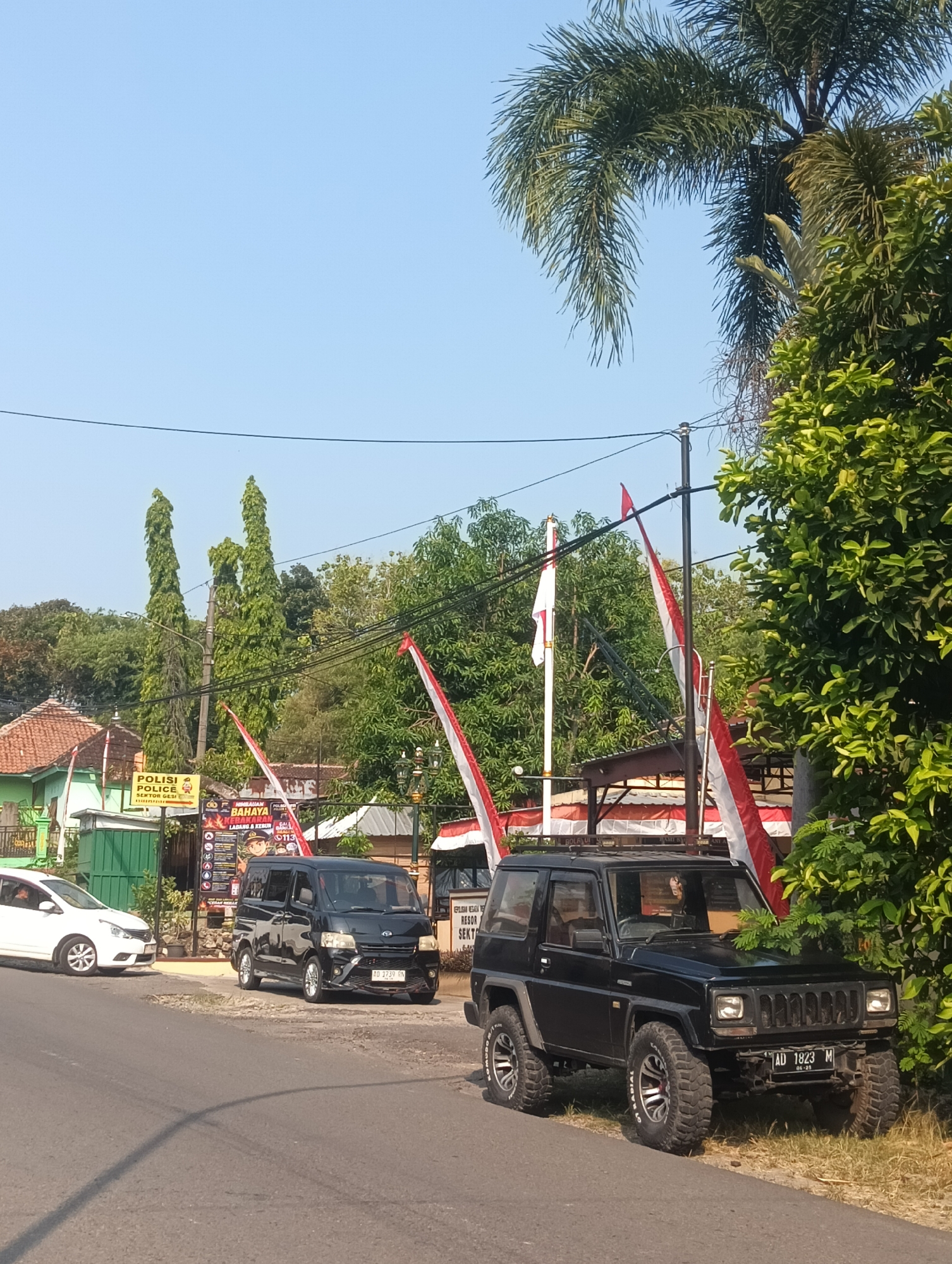 The image depicts a street scene in Palemrejo, Indonesia, on a clear, sunny day. In the foreground, a black, two-door SUV with large off-road tires is parked on the side of the road, facing right. Its license plate reads "AD 1823 M". To the left of the SUV, a black minivan is also parked, facing right, with the license plate "AD 1739 RN". Further to the left, a white car is partially visible, facing left.
Behind the parked vehicles, a building with a red-tiled roof and green walls is visible. A sign with "POLISI POLICE SEKTOR GESI" in yellow lettering is attached to a pole in front of this building. Adjacent to the building, a large banner with Indonesian text and images, including a depiction of a firefighter, is displayed.
Several red and white Indonesian flags are mounted on poles and are fluttering in the breeze. Tall, slender green trees are prominent in the background, along with a large palm tree on the right side of the image. Electrical wires are strung across the sky. The overall environment suggests a public area, possibly a commercial or administrative zone, adorned with national symbols.