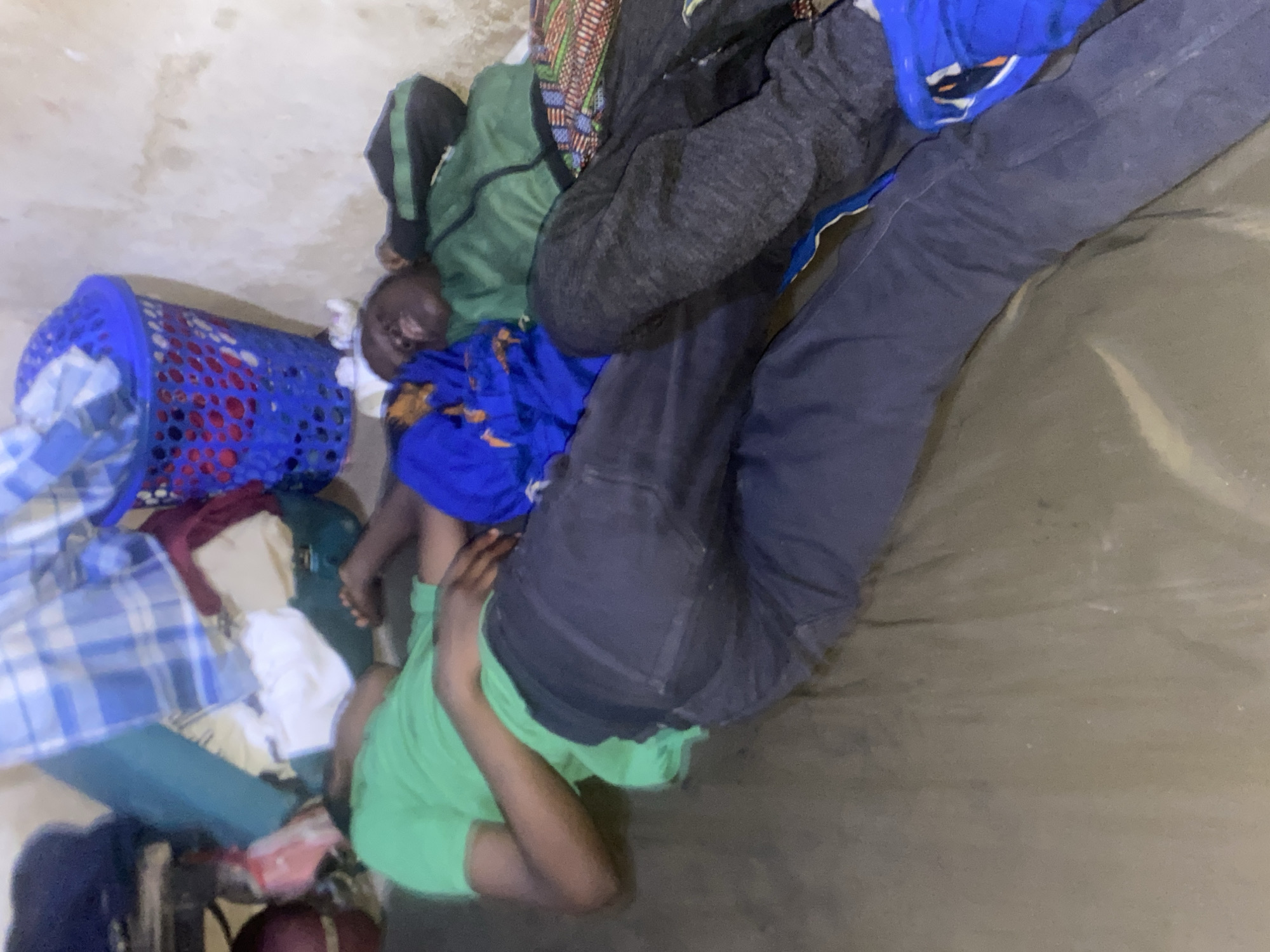 Location: Bukuru, Nigeria
A man in a green shirt is lying on a gray surface. A person wearing jeans and blue shoes is standing near him. A blue laundry basket filled with clothes is next to the man in the green shirt. Other objects include a green jacket, a gray sweatshirt, and various fabrics. The scene appears to be indoors, possibly in a room with a light-colored wall.