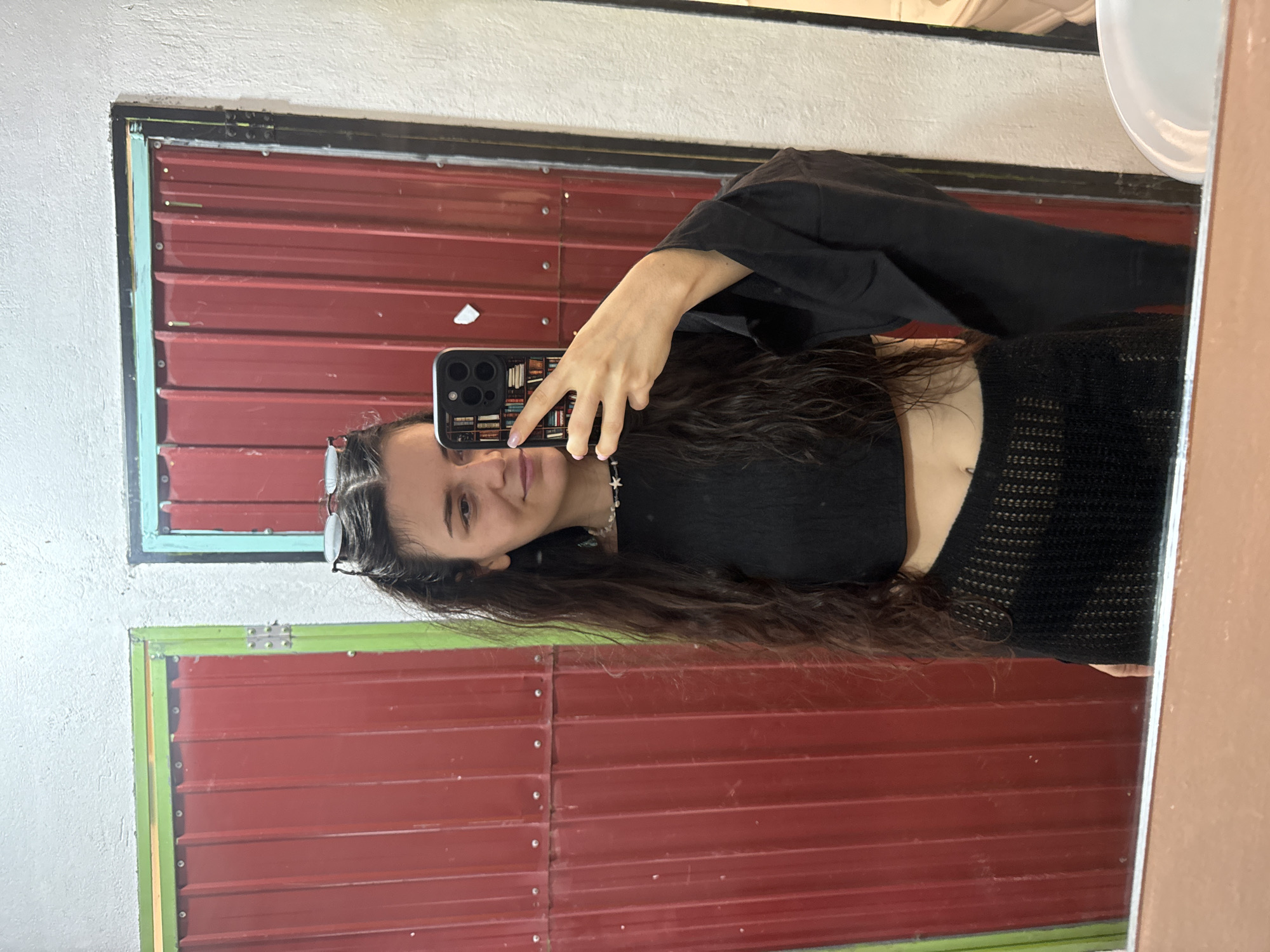 A young woman with long, wavy dark hair is taking a selfie in a mirror. She is wearing a black cropped top and a black cardigan or jacket. The woman is holding her phone with her left hand, displaying a phone case with a book motif. She is also wearing glasses on her head and a delicate necklace.
The mirror frame is visible and shows red horizontal panels with visible seams and rivets. The panels are framed by green and blue trim. The background suggests the setting is likely a utilitarian space, possibly a bathroom, with a white sink partially visible in the upper right corner. The scene is undated, and there is no definitive evidence to confirm that this is located in Mũi Né, Vietnam.