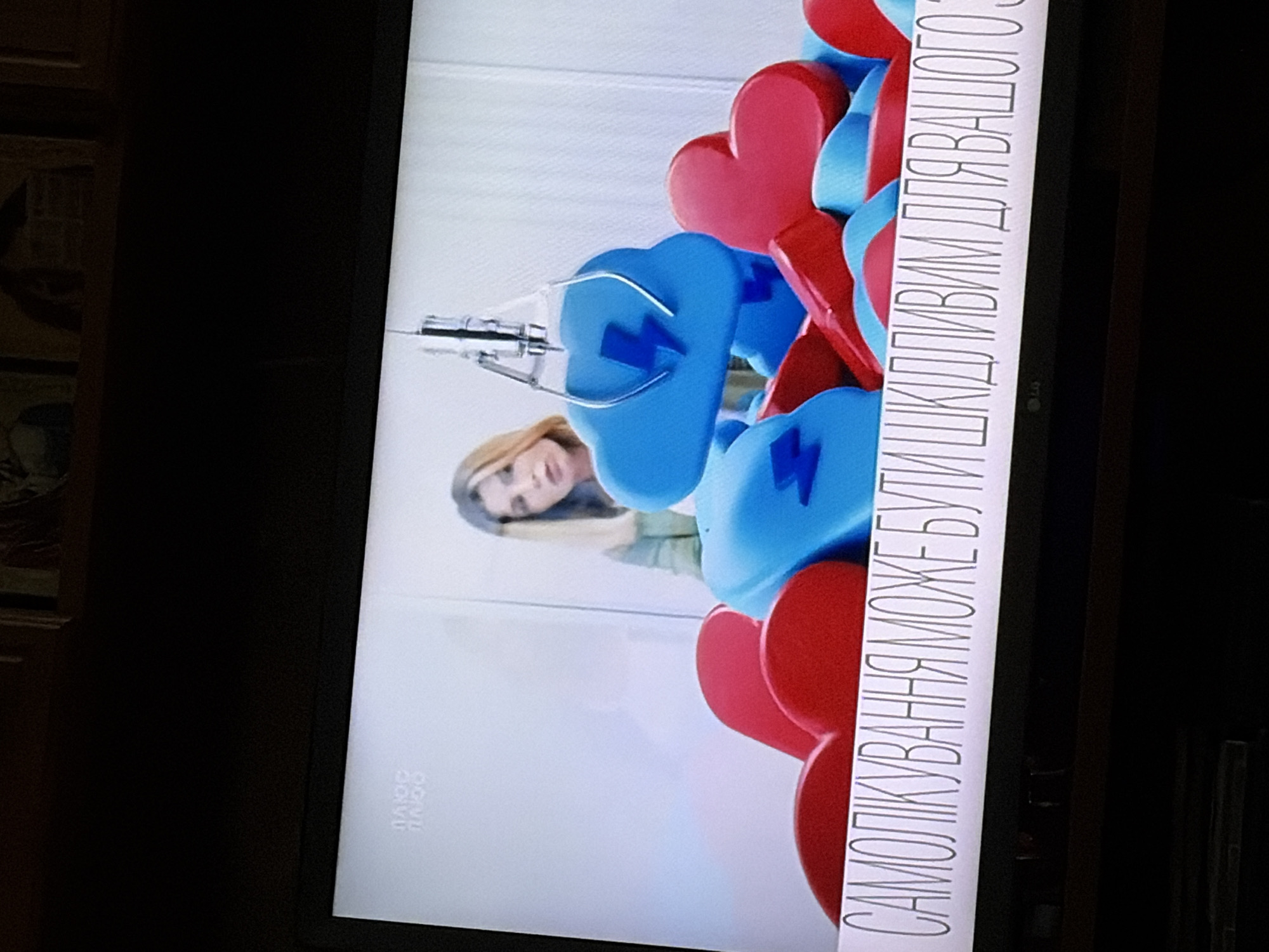 The image shows a television screen displaying a news segment. In the background, a woman with blonde hair is visible behind a pile of red heart-shaped objects and blue cloud-shaped objects, some of which have lightning bolt symbols on them. A claw from a game machine is positioned above the objects, appearing to be picking up a blue cloud. At the bottom of the screen, there is text in Ukrainian that reads "САМОЛІКУВАННЯ МОЖЕ БУТИ ШКІДЛИВИМ ДЛЯ ВАШОГО ЗДОРОВ'Я" which translates to "Self-treatment can be harmful to your health." A small "LG" logo is also visible at the bottom of the screen. The setting is an indoor environment, likely a studio.