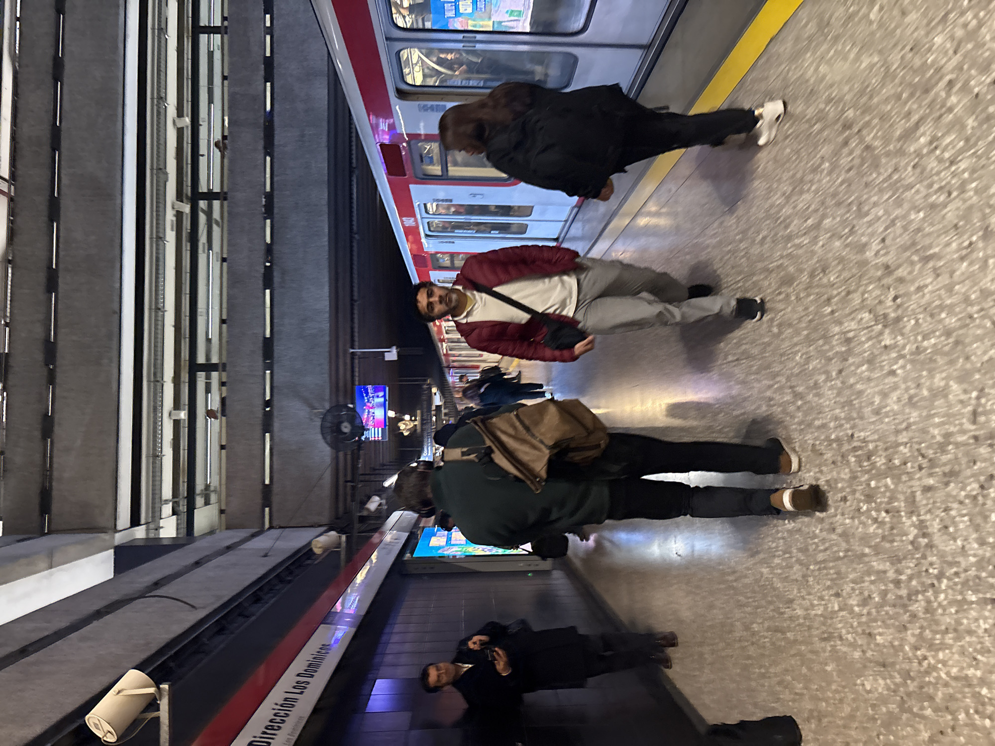 The image shows a subway platform in Santiago, Chile. A train is stopped at the platform, and several people are walking. The platform is made of speckled concrete, and the walls are gray and ribbed. A sign on the wall reads "Dirección Los Dominicos." The train is white with red trim, and its doors are open. A man in a dark coat is standing on the platform, looking at his phone. Another man in a green jacket is walking away from the camera, carrying a brown backpack. A man in a red jacket and gray pants is walking towards the camera, with a black bag slung across his chest. A woman in a black jacket is walking away from the camera. The lighting is artificial, creating shadows on the platform.