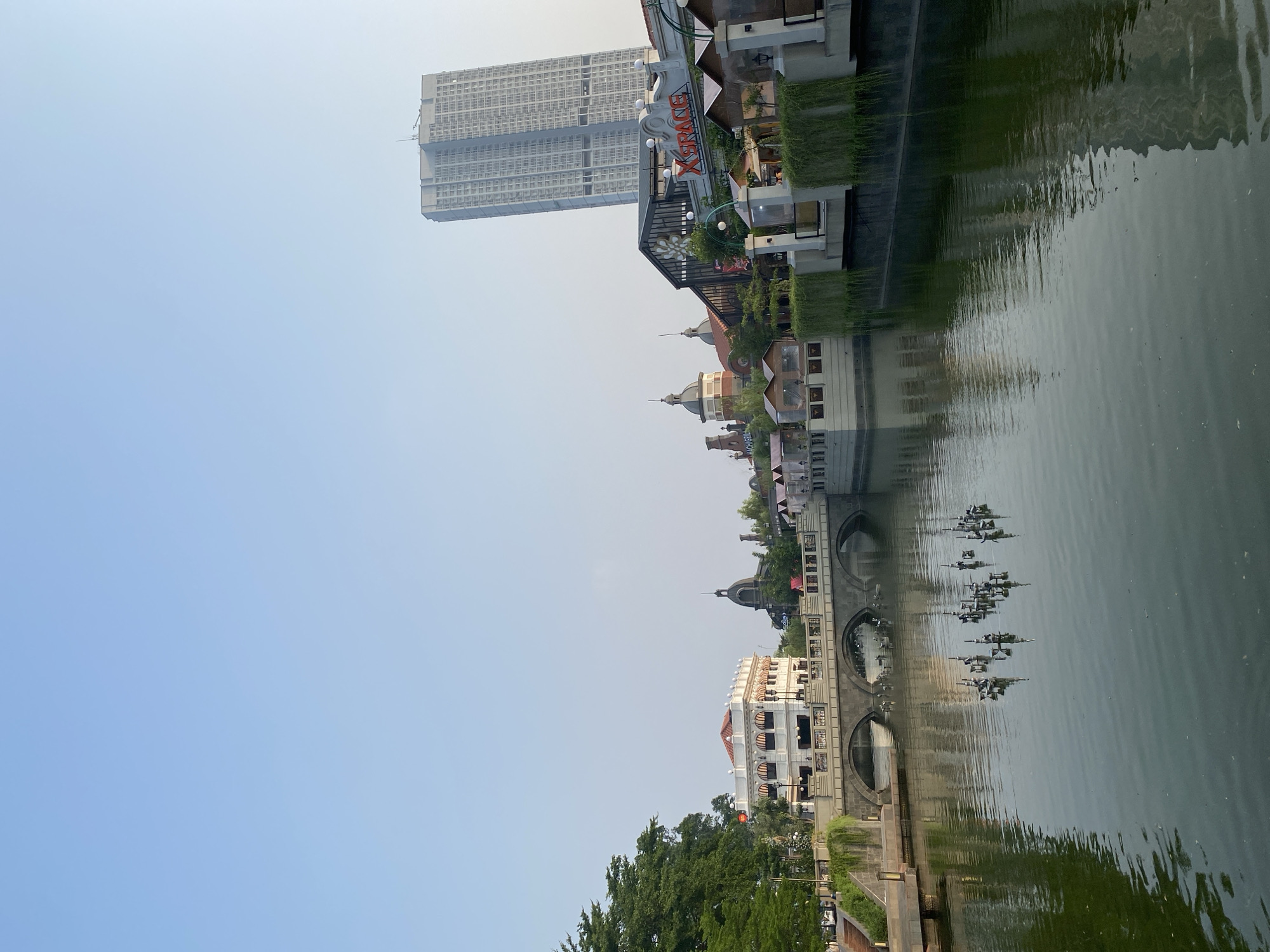 The image shows a waterfront scene with buildings and a tall skyscraper reflected in the water. The buildings have a European architectural style, with domed roofs and arched windows. Some of the buildings are adorned with hanging plants. A large, modern skyscraper stands in the background, its reflection distorted in the rippling water. The sky is a clear, pale blue, suggesting it is daytime, likely morning or late afternoon due to the soft light. The water appears calm, with gentle ripples.
The scene is set in a developed urban area, with the presence of both historical-style architecture and a contemporary skyscraper indicating a mixed-use environment. There are no people visible in the image. The overall mood is tranquil and picturesque, emphasizing the interplay between the built environment and its reflection.
Visible text includes "XGRAND" in orange lettering on a sign above one of the buildings, and another partially visible word "CORPO" on a building facade across the water. The location context is East Jakarta, Indonesia.