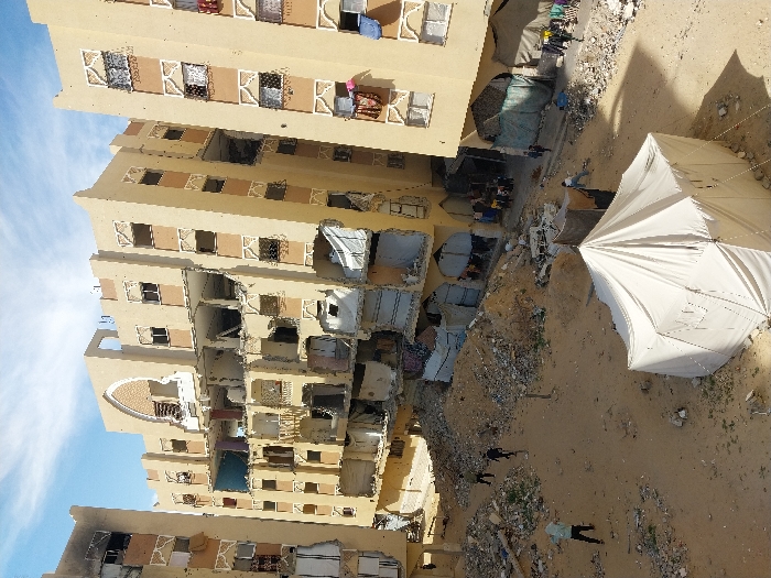 This image depicts a devastated urban landscape in Khan Yunis, Palestinian Territory. The scene is dominated by three multi-story residential buildings. The central building exhibits catastrophic structural damage, with its facade extensively destroyed from multiple floors upwards, exposing numerous interiors and leaving gaping holes where walls and ceilings once stood. The building to its left shows visible damage, particularly on upper floors, with exposed rooms and potential burn marks. The building on the right appears less structurally compromised but has exposed window interiors and some visible debris on balconies.
The ground level is an expanse of sandy earth, heavily strewn with large piles of rubble, concrete fragments, and construction debris, indicative of widespread destruction. In the foreground, a large, white canvas tent is erected on the sandy ground, to the right of the frame's center. Several individuals are present on the ground. One person in a light top and dark pants is walking left-to-right in the mid-ground. Further back, two individuals in dark attire walk towards the left. Near the base of the rightmost building, a small group of people is visible. Another person in a light top is positioned to the left of the tent. Makeshift shelters or tarpaulins are visible near the lower levels of the buildings, and clothes are seen hanging from windows or makeshift lines. The sky is partially cloudy blue, and shadows indicate a daytime setting.