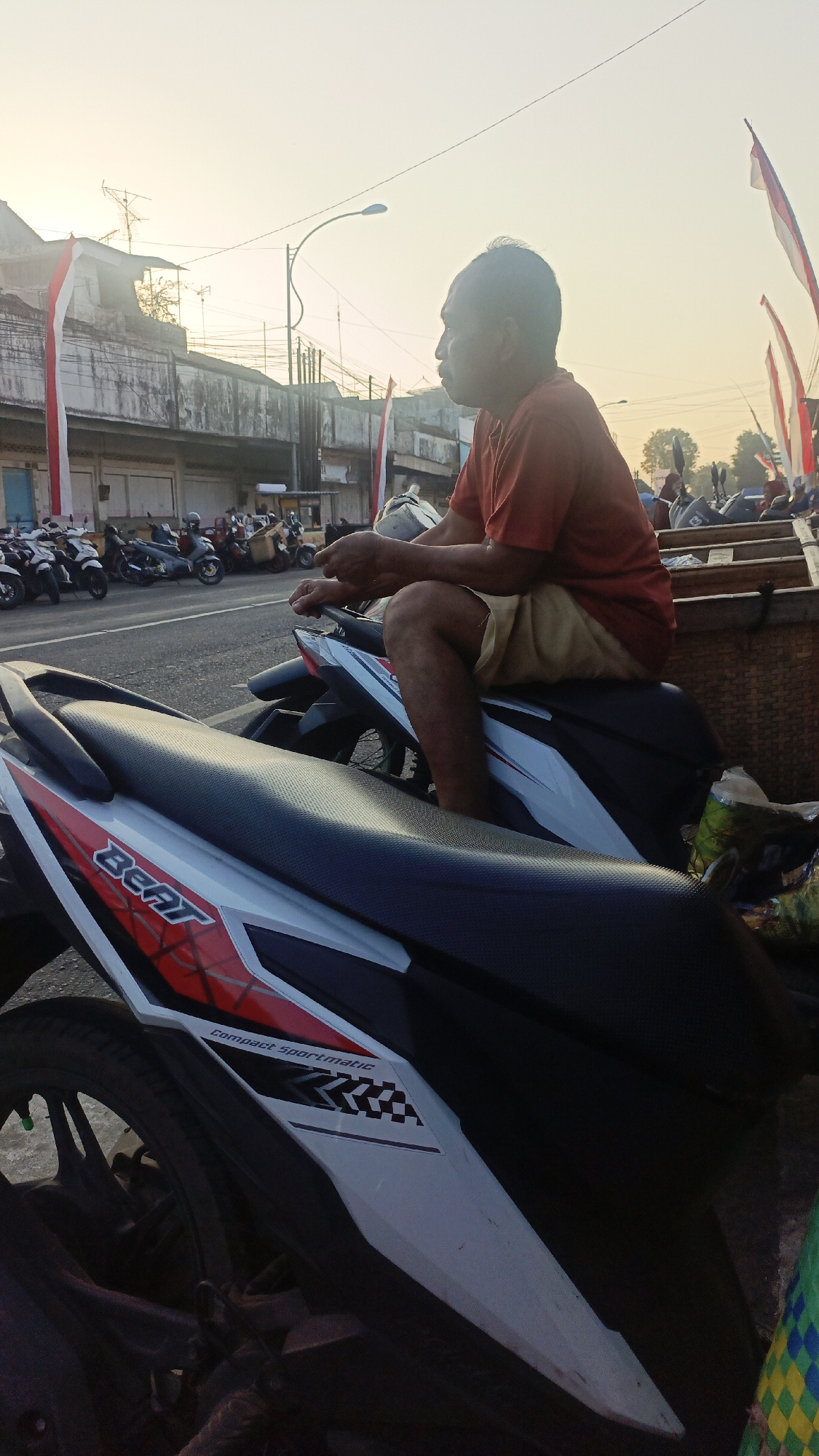 In Srono, Indonesia, a man sits on a motorcycle in the early morning. The sky is a pale yellow, indicating sunrise. The man, wearing a red shirt and tan shorts, is looking to his right. His hands are resting on the handlebars of the motorcycle he is sitting on. Another motorcycle is in the foreground, and several more are parked in the background. The buildings behind the motorcycles are simple, with a faded red and white Indonesian flag hanging from one of them. A street lamp is visible in the distance. The scene appears to be a roadside or parking area.