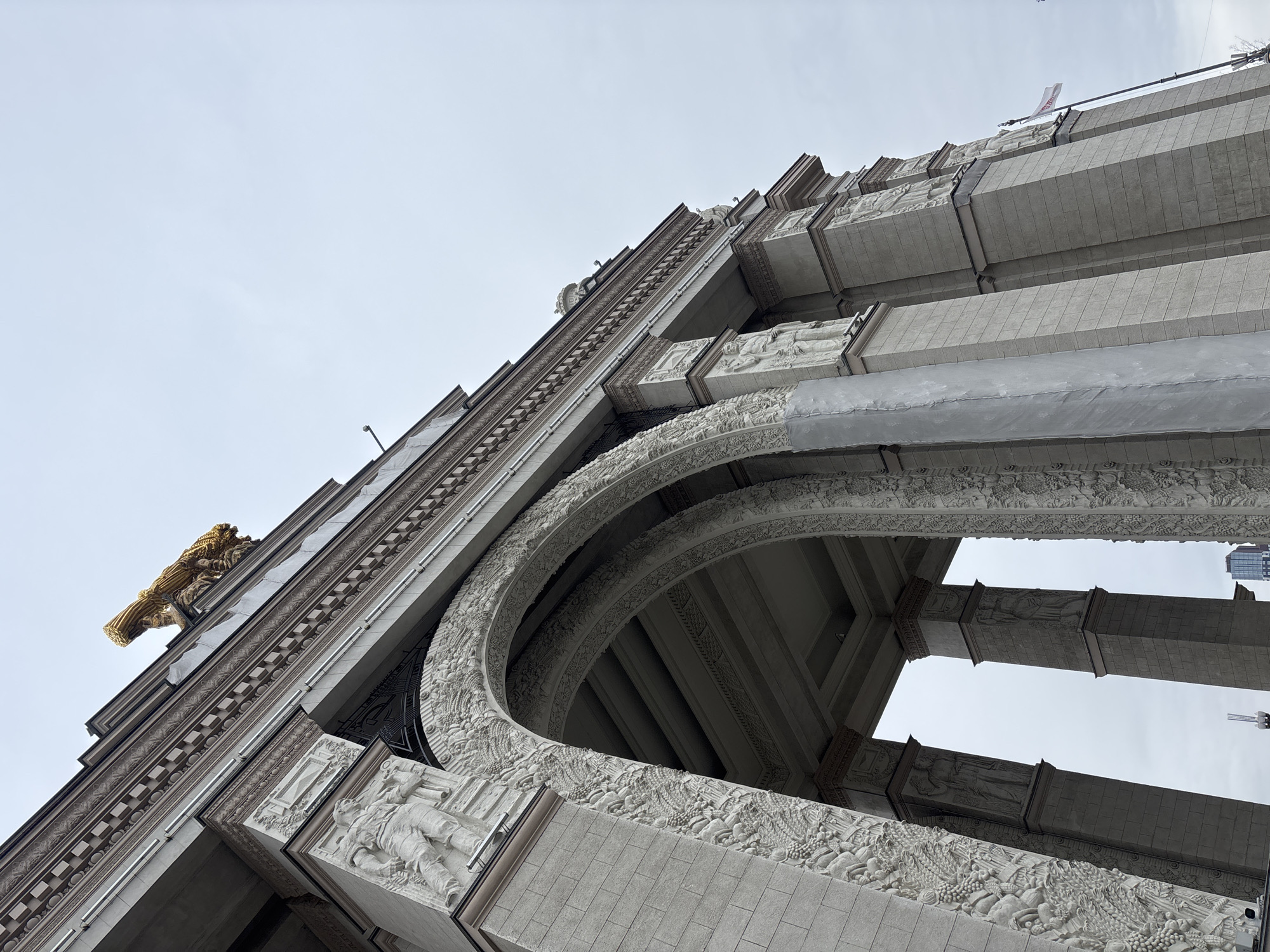 This low-angle photograph showcases a portion of the facade of a monumental building in Moscow, Russia. The architectural style features ornate details and relief sculptures. A golden sculpture of a wheat sheaf with sunflowers and a hammer and sickle is prominently displayed at the top corner. The facade exhibits a series of arches adorned with intricate carvings and detailed depictions of figures. The building's structure is layered with various architectural elements, including cornices and projections. The sky is visible in the background.