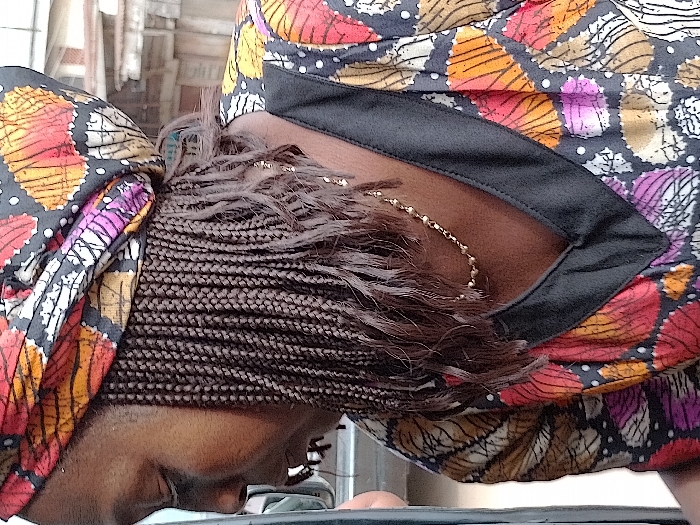 A person with dark skin and braided hair adorned with a colorful headscarf bends forward. The headscarf features a mix of orange, red, pink, and white patterns. The person wears a blouse with a similar colorful pattern and a black collar. A delicate gold necklace is visible at the base of the neck, peeking out from beneath the hair. The focus is on the back of the head and neck, partially obscured by the braided hair.
