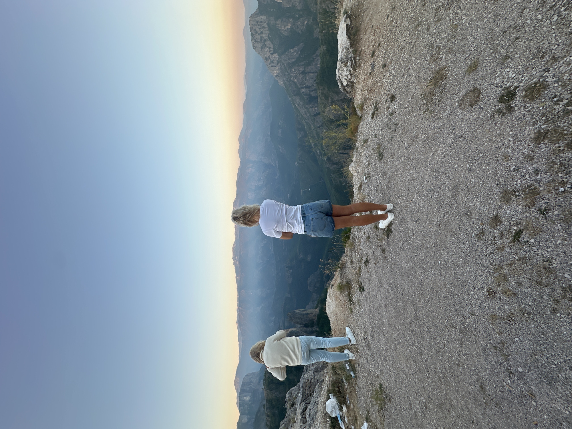 Two individuals are standing on a rocky cliff overlooking a vast mountain landscape at dusk. The person on the left, facing away from the camera, is wearing a light-colored sweater and light blue jeans. The person on the right, also facing away, is wearing a white t-shirt and denim shorts. Both individuals are looking out at the mountainous vista, with the sky showing a gradient of blue and orange hues. The terrain in the foreground is covered with small stones and sparse vegetation. In the distance, layers of mountains recede into a hazy, blue-toned valley. There is also some litter visible on the cliff edge near the left individual. The scene captures a moment of quiet contemplation of nature.