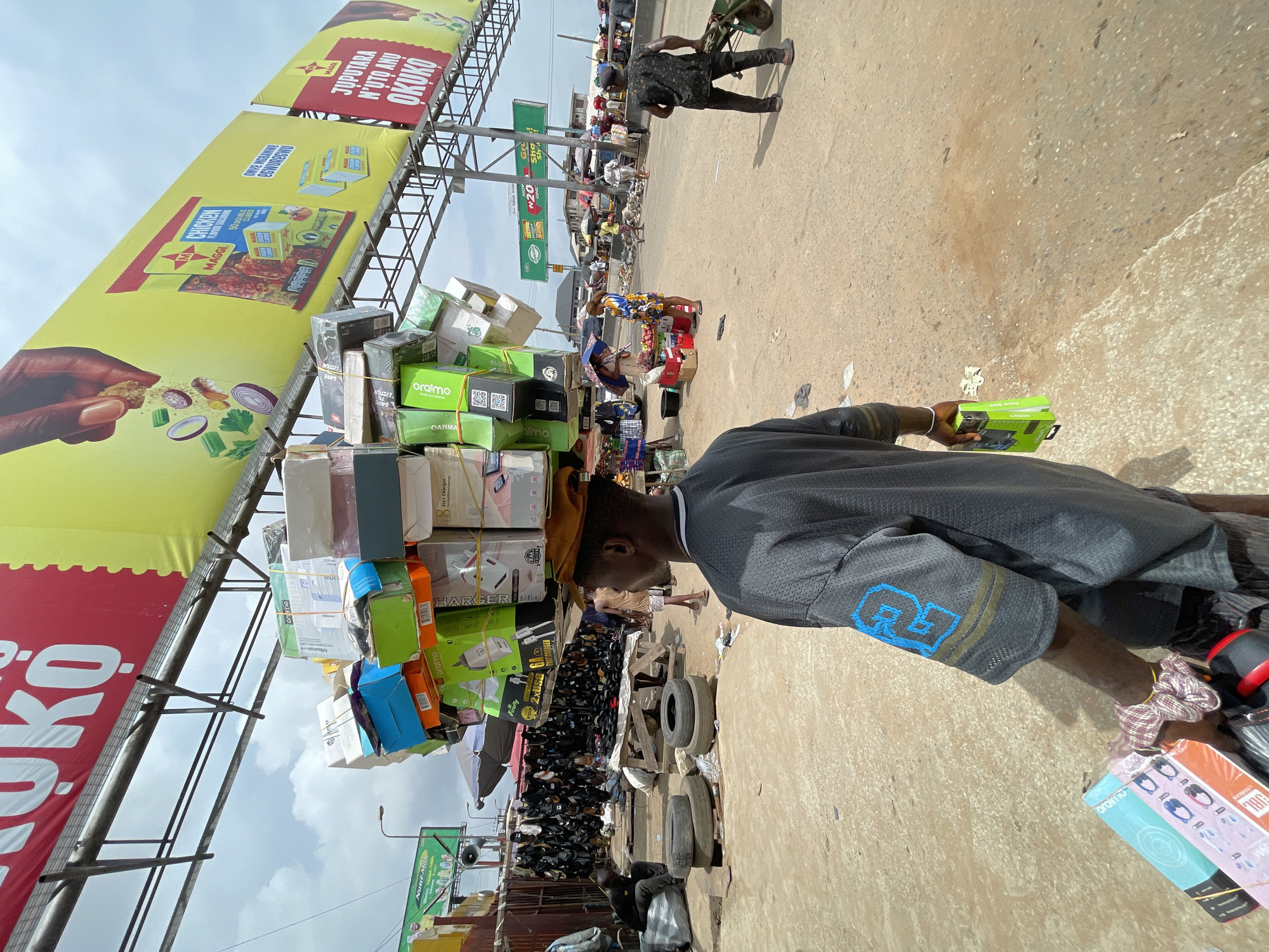 The image shows a bustling marketplace in Onitsha, Nigeria. A man is carrying a large assortment of goods, primarily electronics like chargers and speakers, balanced on his head. He is wearing a gray shirt with the number "23" on the sleeve. In the background, vendors and shoppers can be seen amongst stalls with various items. Above the scene, large billboards advertise products such as Maggi bouillon cubes. The ground is paved with what appears to be a mix of concrete and dirt. Car tires lean in the background.