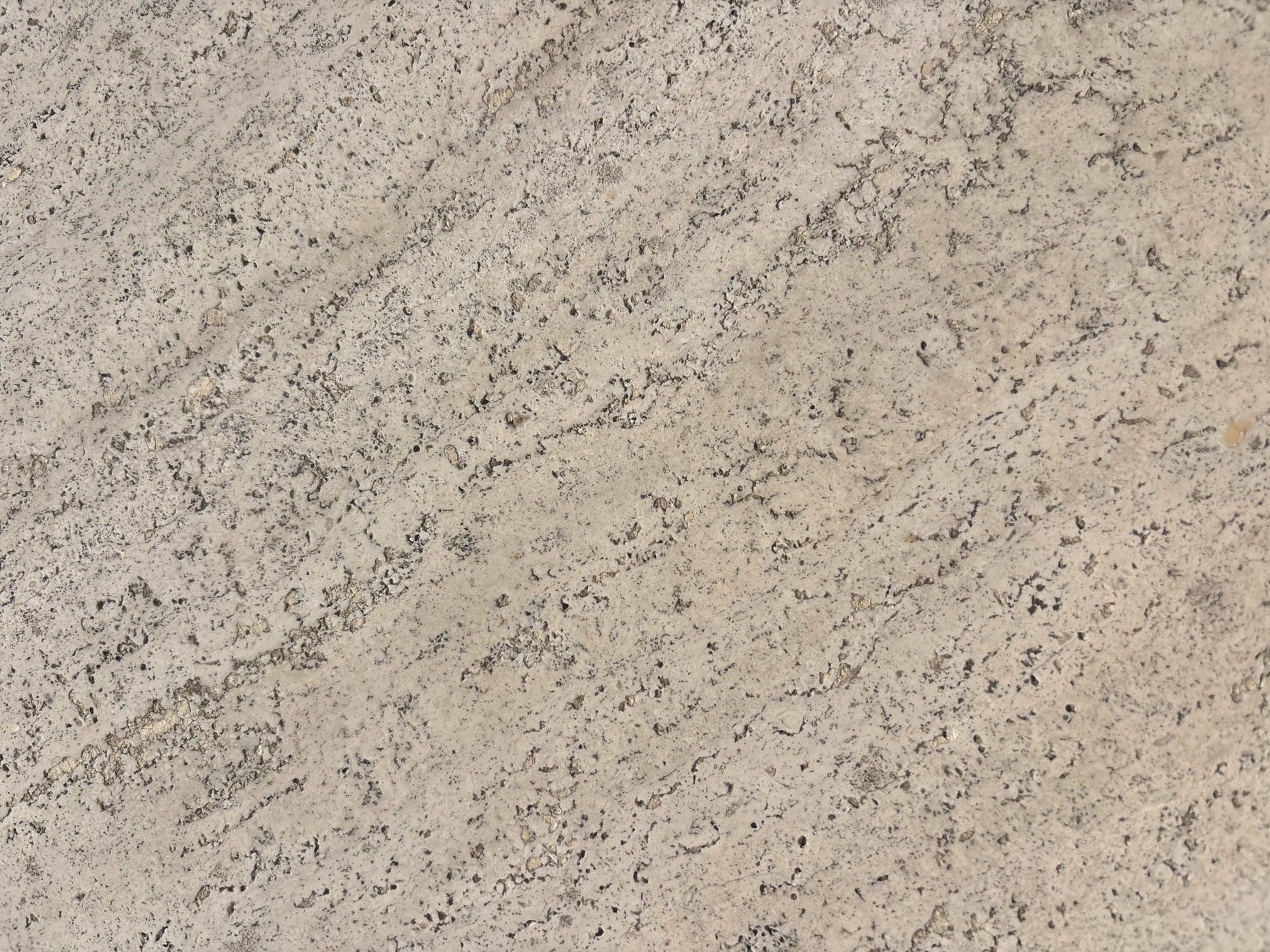 The image shows a close-up of a textured stone surface. The stone is predominantly light beige or off-white with dark grey or black speckles and small grooves. The pattern is linear, running from the top left to the bottom right of the image. It has a rough and porous texture. It may be of travertine stone, which is common in Rome, Italy. However, without additional information, the precise location and origin of the stone cannot be determined.