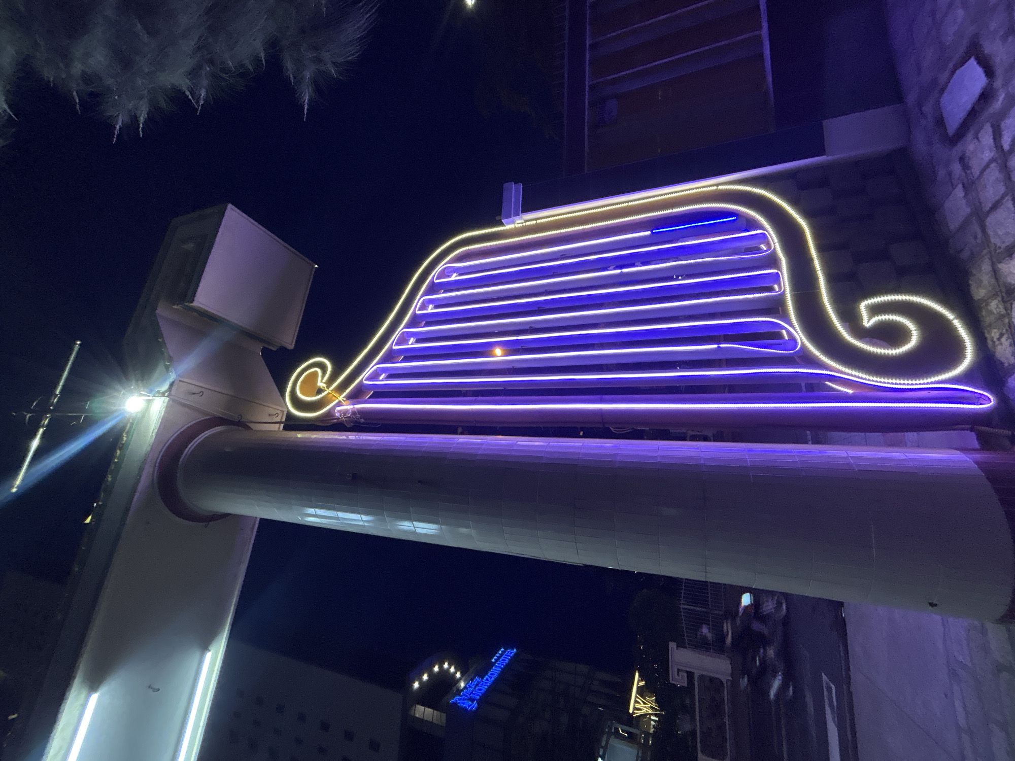 The image shows an upward shot at night in Bac Nha Trang Ward, Vietnam. A large, brightly lit architectural structure dominates the foreground, resembling a fedora hat outlined with gold and filled with horizontal blue neon lights. This structure is mounted atop a cylindrical pillar, also illuminated, with a white and silver support. Below the pillar, in the background, the "Horizon Hotel" sign is visible in blue neon, along with other illuminated buildings, suggesting an urban setting. The surrounding environment is dark, with hints of vegetation overhead.
