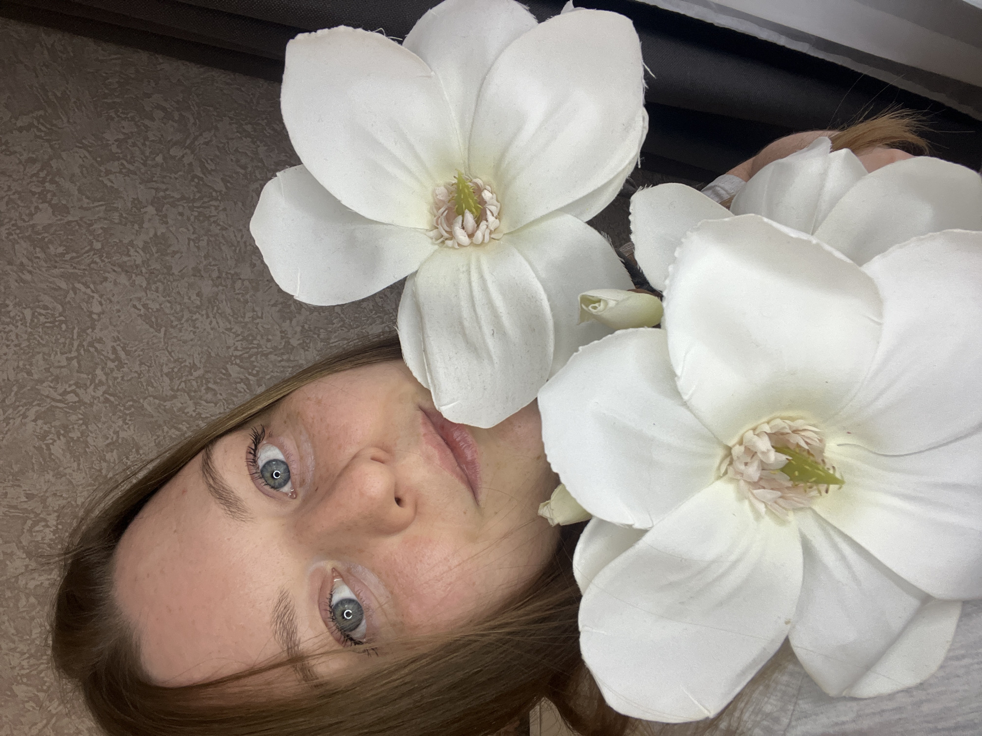 A light-skinned woman with freckles and blue eyes is centrally framed with two large white artificial magnolia flowers positioned at the top-left and bottom-right of the frame. The woman's face is slightly rotated to the left, and her hair appears to be light brown. The background is a textured surface, possibly wallpaper, in muted earth tones. A dark colored curtain partially covers the top of the frame. The subject's gaze is directed upward toward the viewer, creating an intimate connection. There is no indication that this image was taken in the city of Razdolie, Russia, or any other location.