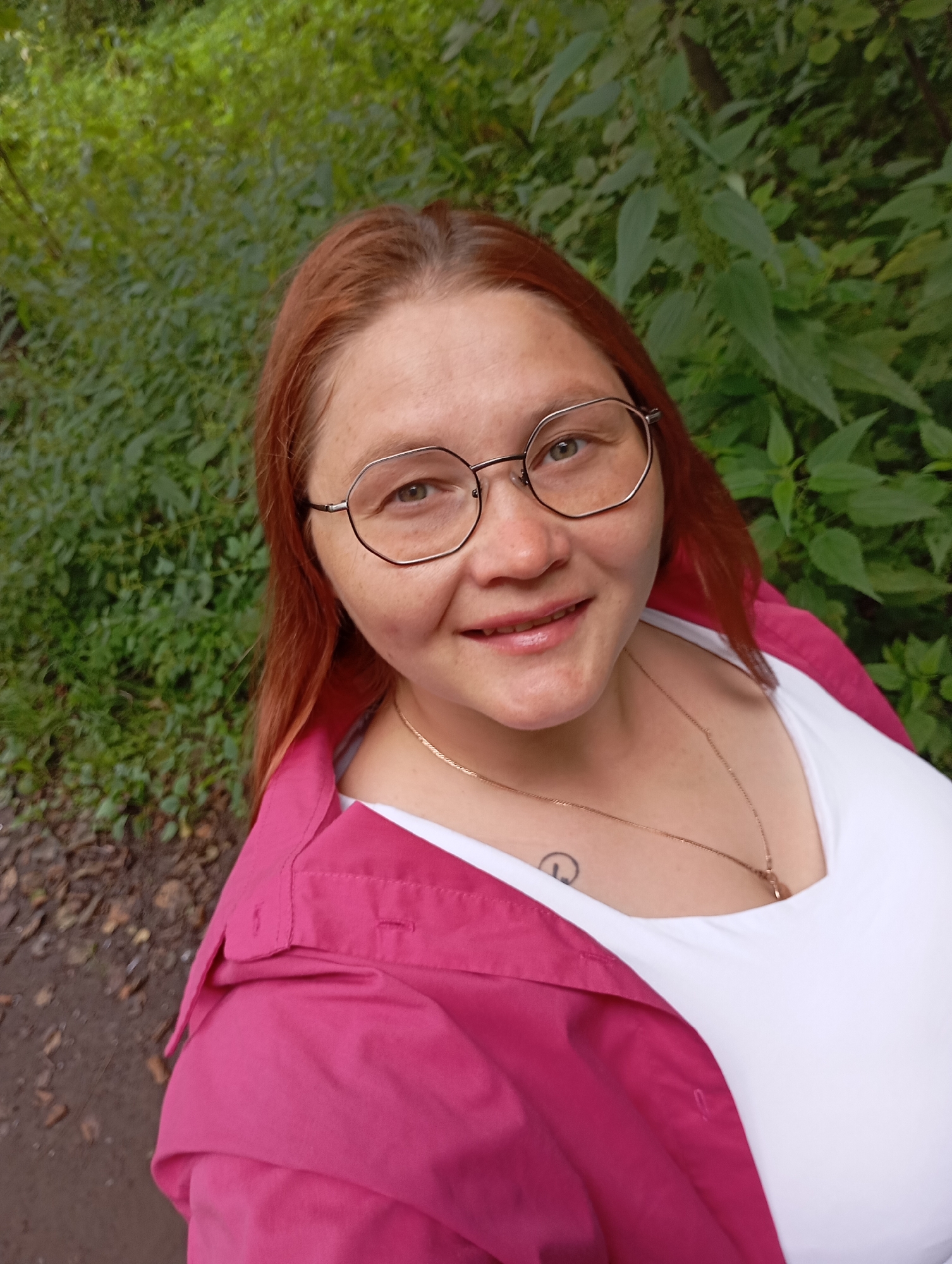 A woman with auburn hair and glasses smiles at the camera. She is wearing a white top and a pink jacket. A gold necklace with a pendant is visible on her chest, as well as a tattoo on her collarbone. The background is a lush green forest with trees and foliage, suggesting a natural outdoor setting. Based on the presence of trees and foliage, this image may have been taken in the city of Chaikovsky, Russia, which is known for its natural beauty and surrounding forests.