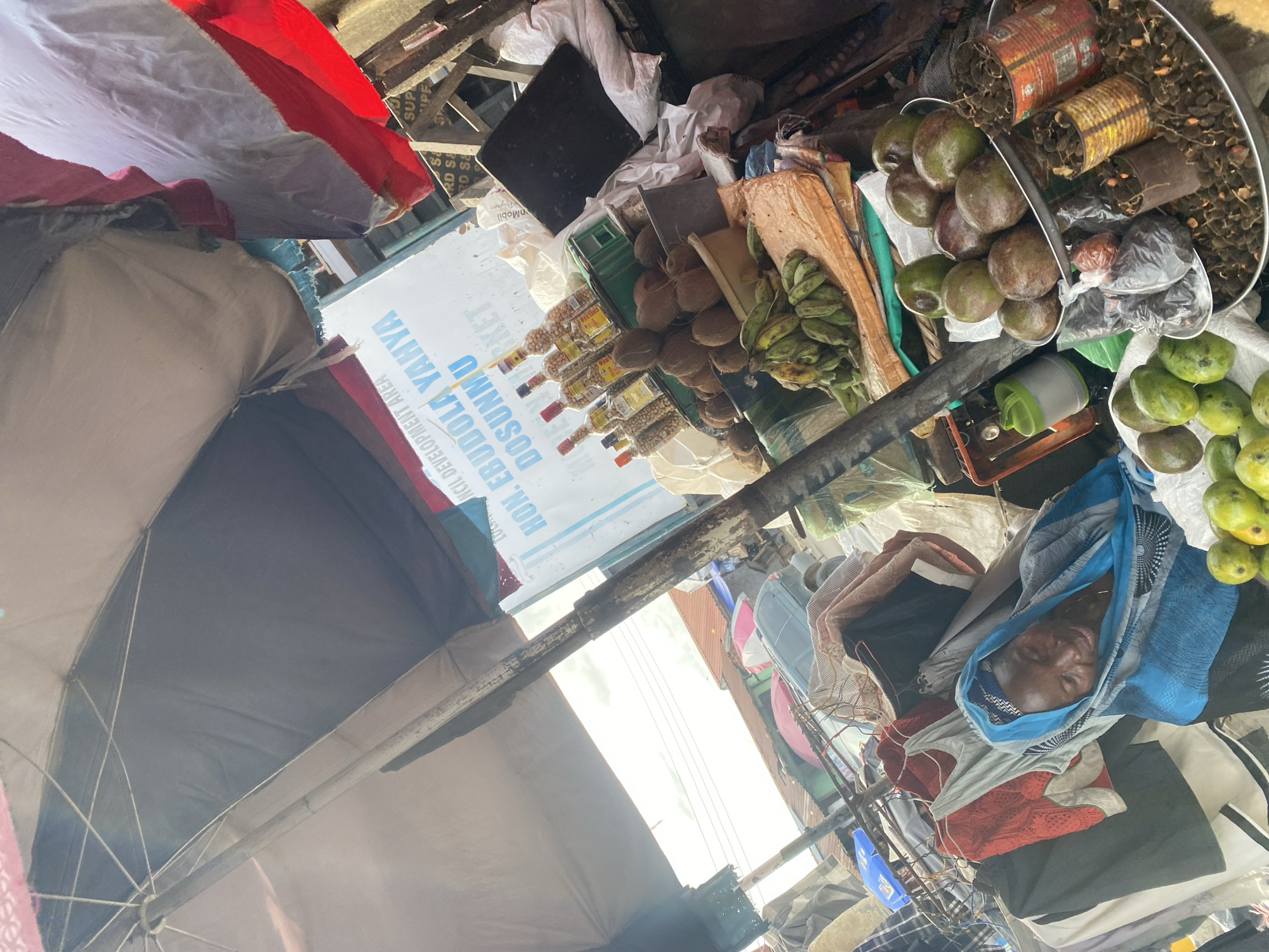 The image displays a lively outdoor market scene, likely in Ado, Nigeria, characterized by an abundance of goods and a relaxed atmosphere. The photo is rotated 90 degrees counter-clockwise, showing the scene from a slightly elevated perspective.
In the foreground, a person is seen resting or lying down, covered partially by a blue and white patterned cloth or blanket, with a calm, almost smiling expression. They appear to be situated amidst various market wares.
The market stall showcases a diverse selection of produce. Prominently featured are large quantities of green mangoes and avocados, piled in metal bowls and a white sack. Bunches of green bananas hang alongside stacked coconuts. Several clear bottles filled with what appears to be peanuts or small seeds are neatly arranged on a counter. Other items include plastic bags of various contents, tin cans, and fabric materials.
The setting is an open-air market, with an umbrella or canopy structure providing shade overhead, its supporting beams and fabric visible. Bright natural light filters into the space, indicating it is daytime. The overall environment appears active yet somewhat cluttered, typical of a bustling local market.
Visible text on signs in the background includes "HON. EBUDOLA YAHYA DOSUNMU" and "INCIL DEVELOPMENT AREA," possibly referencing a local dignitary or community development initiative. Other fragmented texts like "RKET" (likely part of "MARKET"), "nMobil," and "SUP" are also discernible, contributing to the authentic market ambiance.