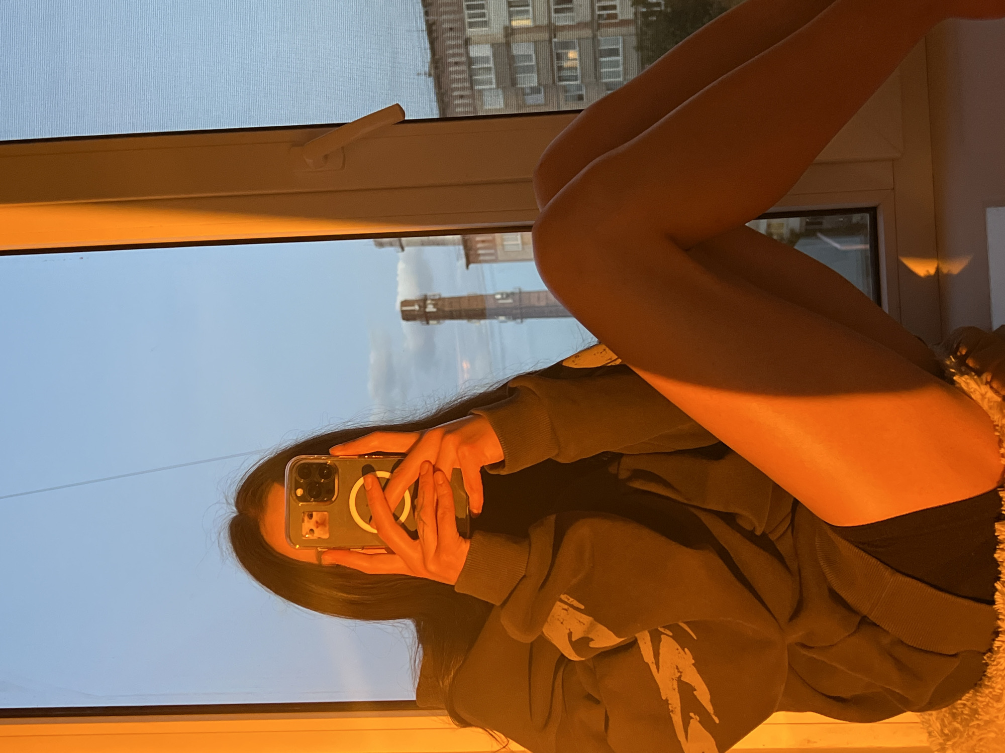 A person with dark hair is sitting cross-legged in a window, taking a selfie. The person is wearing a dark-colored sweatshirt with a white design on it. Their legs are bare. The phone case is clear with a white circle on the back and a small sticker of a cat. Outside the window, a building with many windows is visible, along with a tall industrial chimney. The photo seems to be taken at dusk or dawn, with soft, warm light. Based on the chimney stack, the location may be in Ternopil, Ukraine.