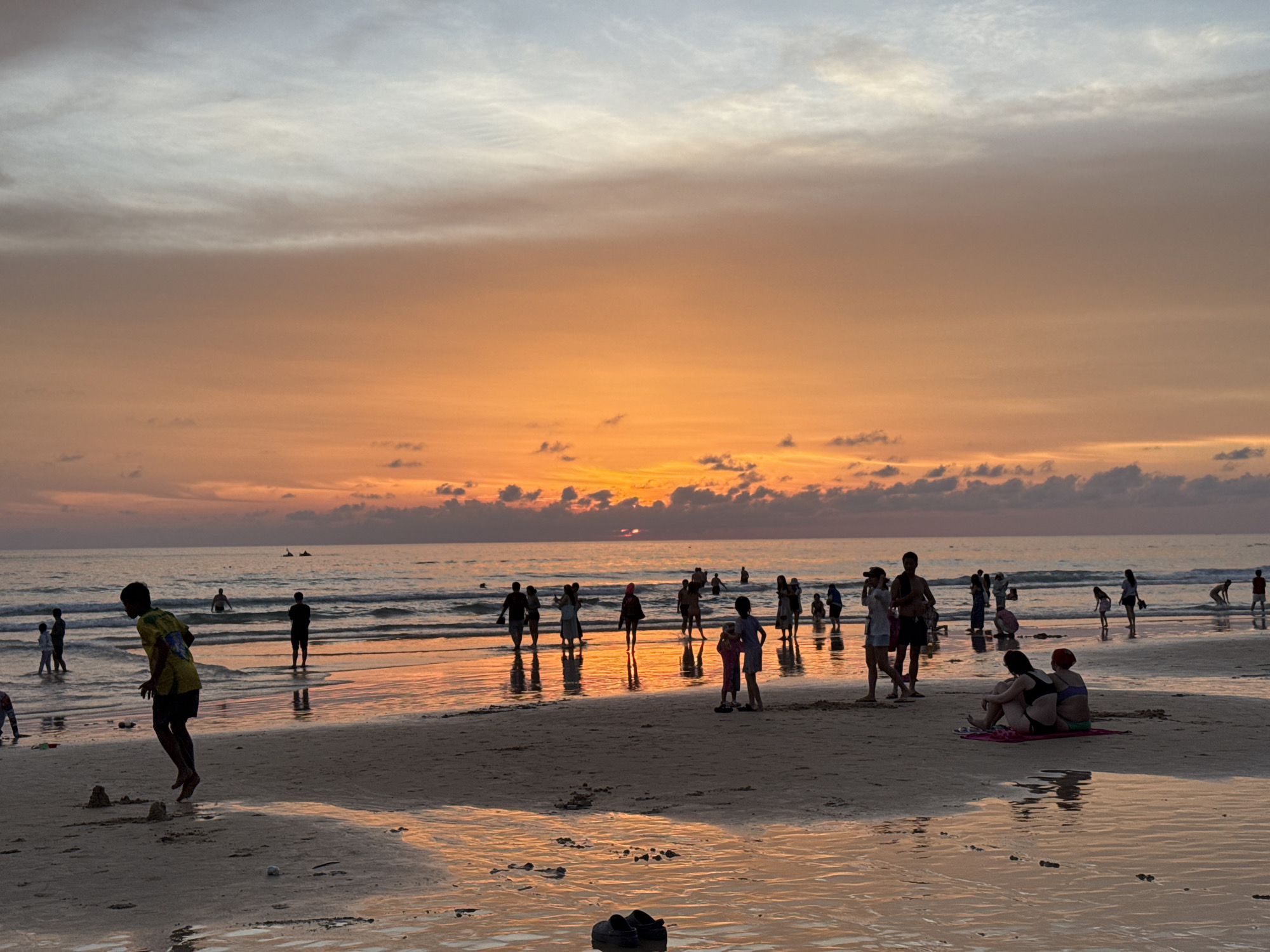 The image depicts a beach scene at Karon Beach, Thailand, during sunset. The sky is a gradient of orange and white, with scattered clouds. The sun is low on the horizon, casting a warm glow on the scene.
Numerous people are present on the beach and in the shallow water. Several individuals are walking along the shore, while others are wading or swimming in the ocean. In the foreground, a young person in a yellow shirt and dark shorts is jumping on the sand. Further back, a group of people are gathered, some standing and others sitting on the sand. Two women are seen sitting on a mat on the beach, facing away from the camera.
The sand is wet in places, reflecting the colorful sky. The ocean appears calm with gentle waves. In the distance, several jet skis are visible on the water. The overall atmosphere is one of a relaxed evening at a popular tourist destination.
