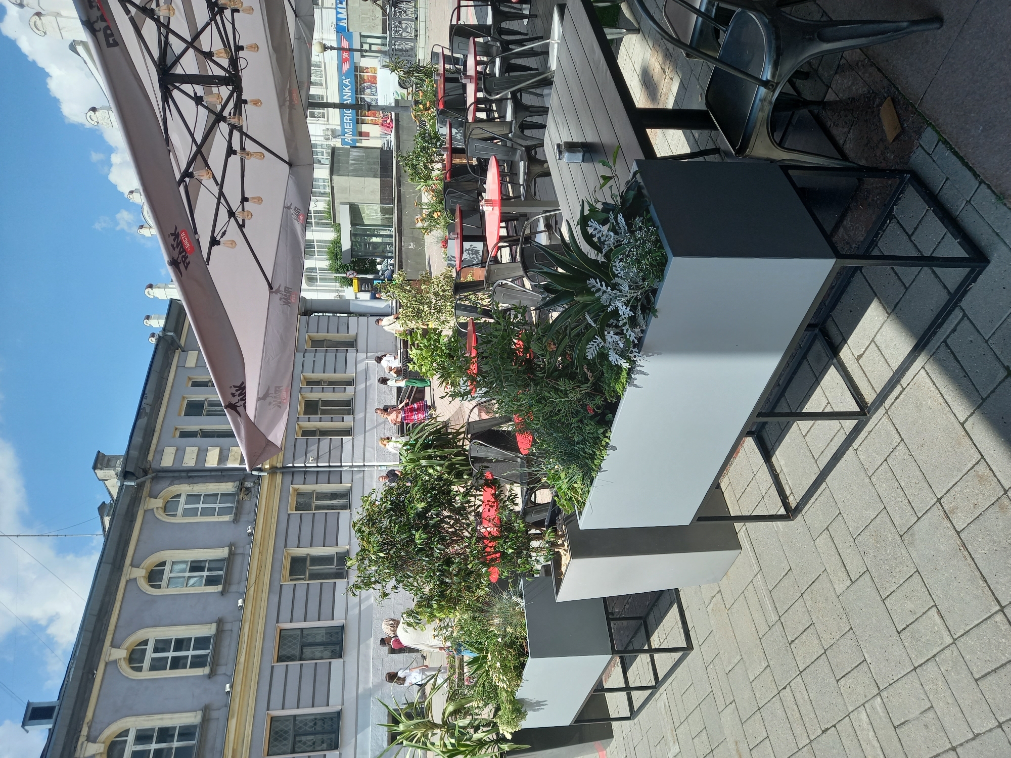 The image shows an outdoor seating area of a cafe in Yekaterinburg, Russia, likely on a sunny day. In the foreground, there are several large, modern grey planters filled with a variety of greenery. These planters are supported by black metal frames, and they are positioned on a light grey brick-paved sidewalk. Behind the planters, tables and chairs for the cafe are visible. Some of the tables are round and red, while others are rectangular and dark grey. The chairs are dark metal with a distressed finish. A large, light-colored umbrella, adorned with lights, covers a portion of the seating area. In the background, a multi-story building with a light grey facade and arched windows is visible. People are walking in the background, suggesting the area is public. The overall impression is a well-maintained outdoor cafe space in an urban environment.