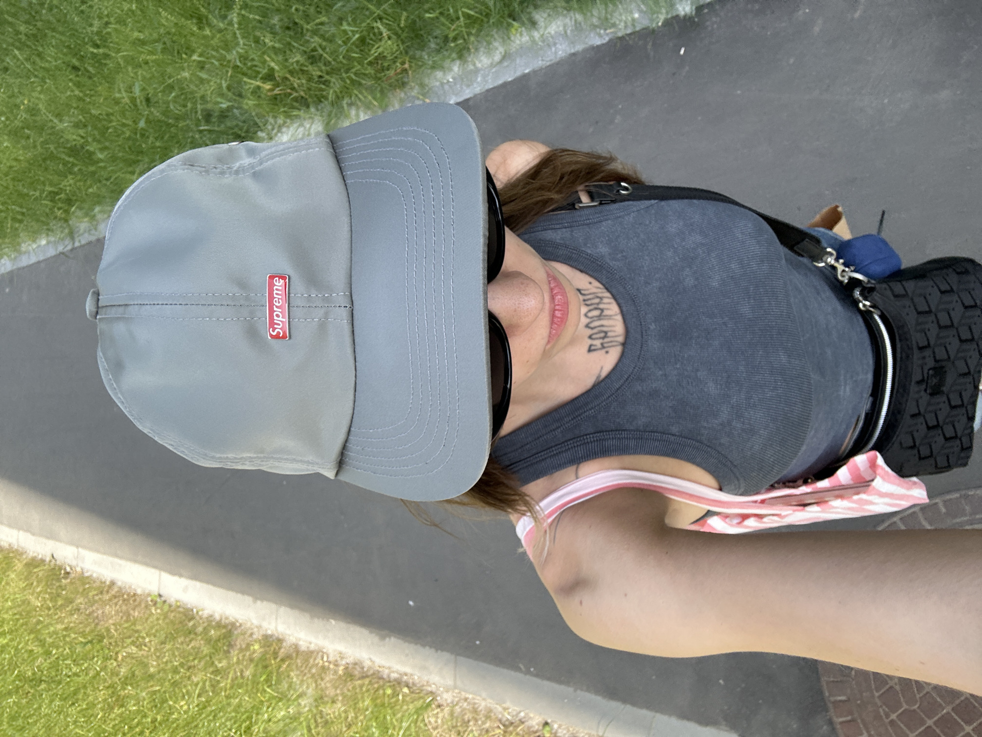 An outdoor, high-angle, point-of-view selfie shows a person from the shoulders up, wearing a gray baseball cap with a red Supreme logo. They are also wearing sunglasses and a gray tank top with a pink and white striped strap visible over their shoulder. A black fanny pack with a textured pattern is worn around their waist, and a small blue object is attached to it. The person has tattoos on their neck and arm. They are standing on a paved path next to a grassy area. The path appears to be a sidewalk or walkway within a park-like setting, possibly in Moscow, Russia, given the context provided. The lighting suggests it is daytime.
