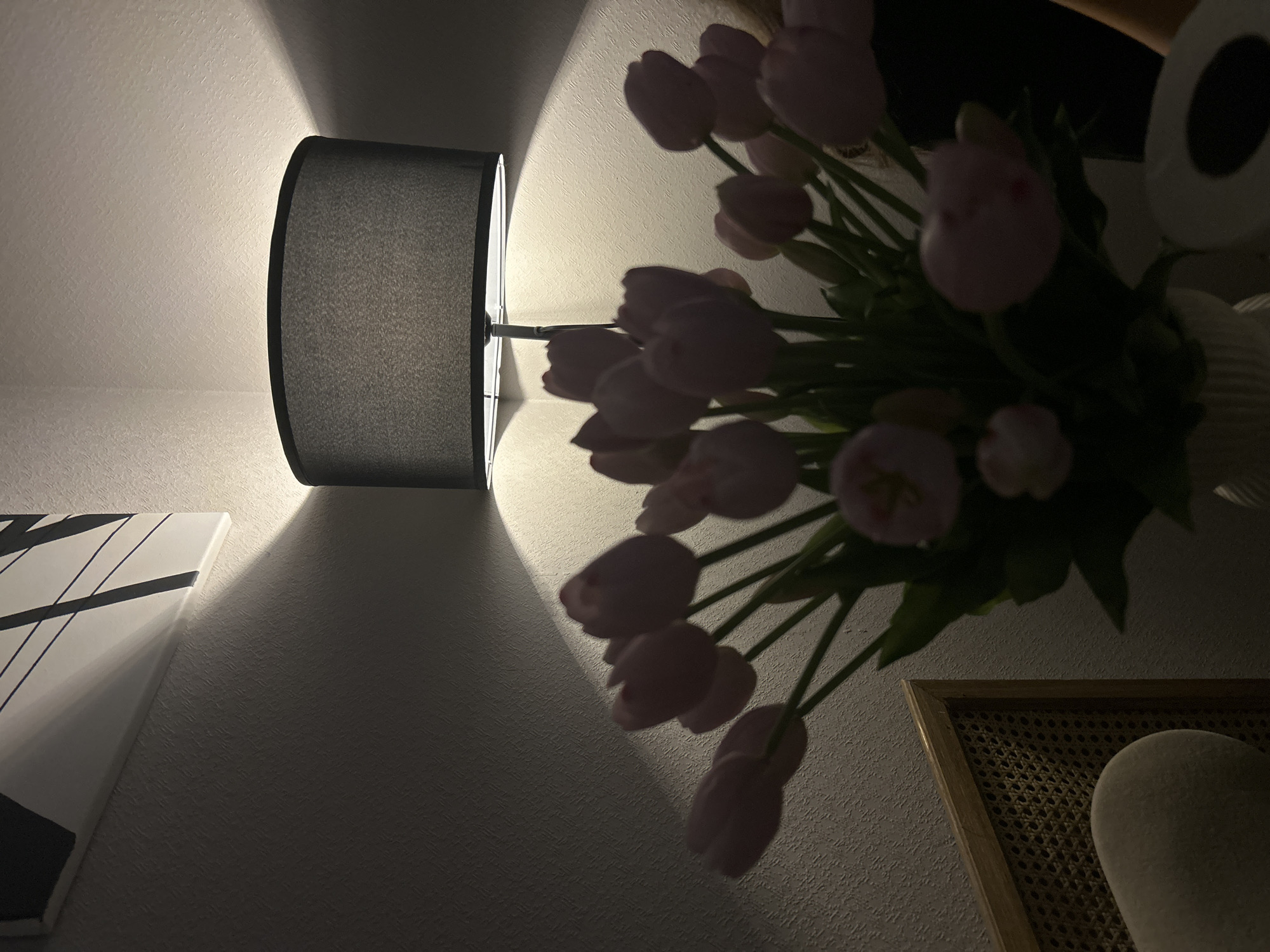 The image depicts a dimly lit indoor scene, possibly a bedroom or living room, with a focus on a bouquet of pink tulips. A black lampshade is visible, casting a warm glow on the textured white wall behind it, creating soft shadows. To the left, a portion of a framed abstract artwork with black lines on a white background is present, also illuminated by the lamp. In the foreground, to the left, a section of a cane-backed chair is visible. The overall atmosphere is serene and intimate. There is no indication that this scene is located in Moscow, Russia.