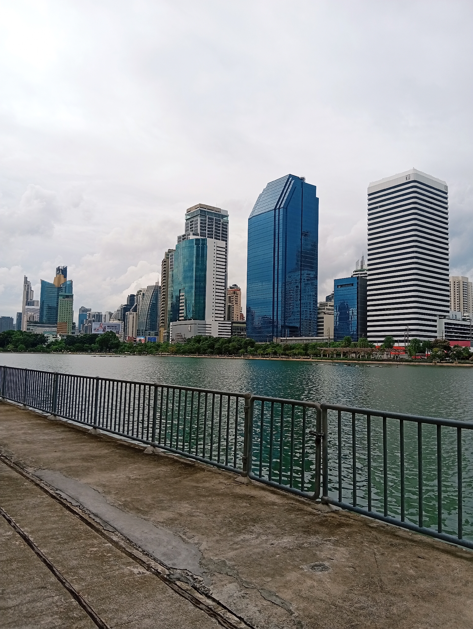 This is a vertical, eye-level, full-shot photograph of the Benjakitti Park in Bangkok, Thailand, captured on a cloudy day. In the foreground, a concrete walkway with visible expansion joints and patches of repair leads toward a dark metal railing. Beyond the railing, a body of water, likely a reservoir, reflects the overcast sky and the buildings in the background. The water has a greenish-blue hue with visible ripples. The middle ground is dominated by a dense line of trees bordering the park. In the background, a panoramic view of Bangkok's modern skyline unfolds, featuring a cluster of tall skyscrapers of varying architectural styles and colors. Notably, a prominent blue glass skyscraper stands out, as does a white building with horizontal black stripes. The sky is overcast with a uniform layer of gray clouds, suggesting a humid or potentially rainy climate. No people are visible in the frame. The overall impression is one of urban development juxtaposed with a man-made green space.
