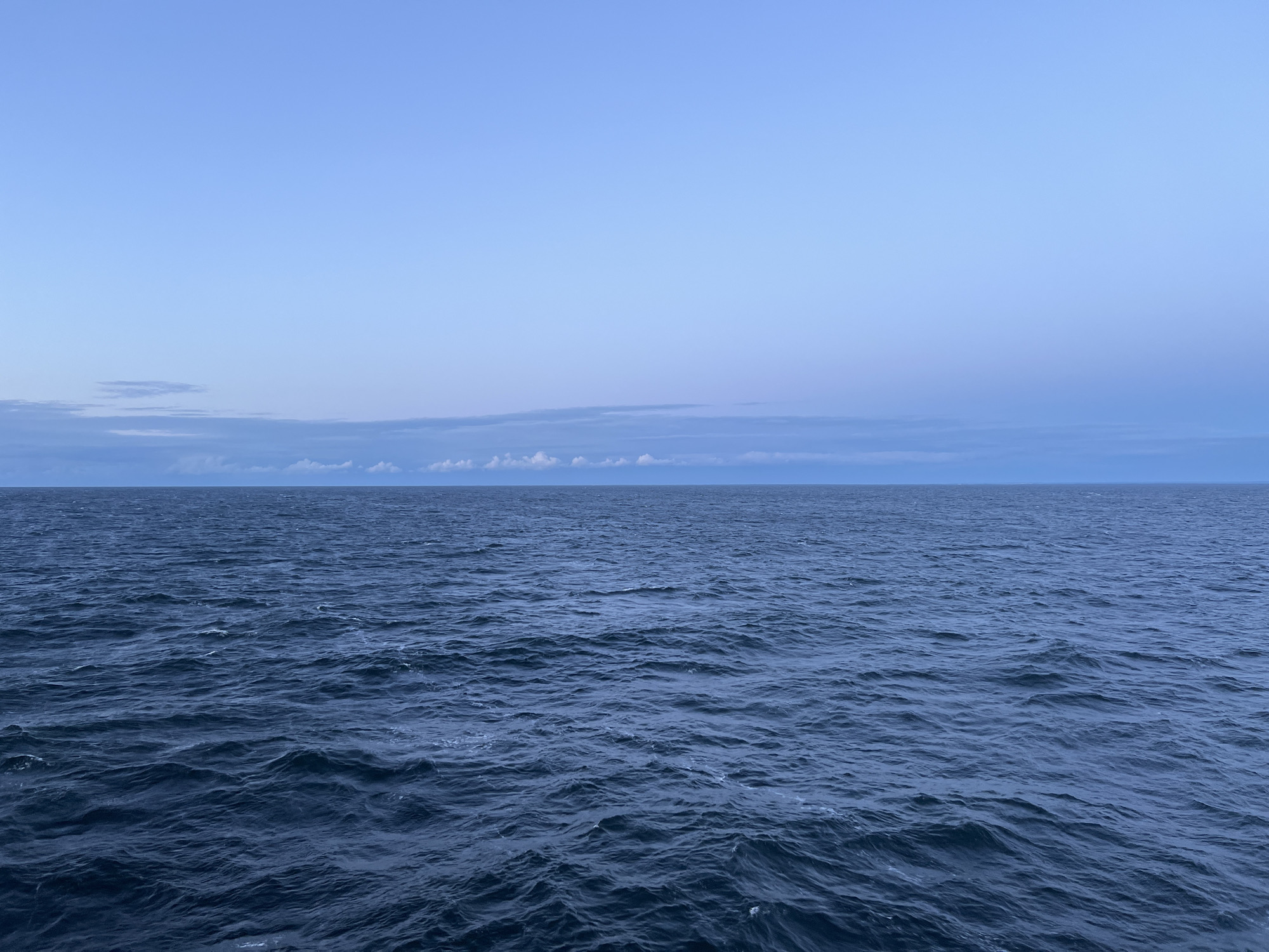 The image displays a vast expanse of dark blue ocean under a pale blue sky. The horizon line, where the sea meets the sky, is clearly defined. Small, white-capped waves are visible on the surface of the water, indicating a moderate chop. A thin band of clouds stretches across the horizon. The scene is devoid of any human presence or man-made structures. The overall impression is one of open water and a clear, cool atmosphere, typical of a coastal or open-sea environment. The location is identified as Unknown, Poland, suggesting a geographical reference point rather than a specific urban area within the city.