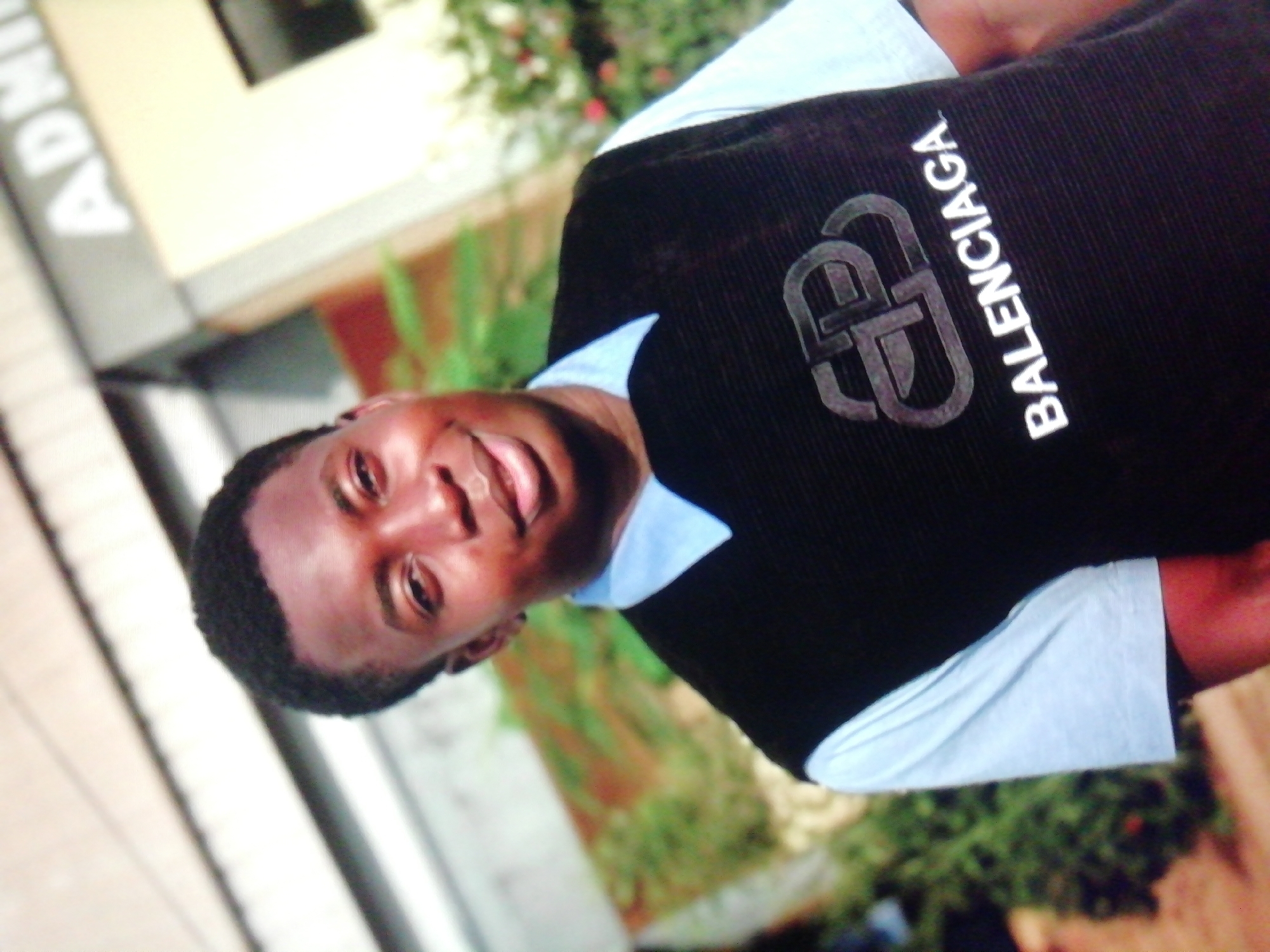 A medium shot depicts a smiling dark-skinned man in a black vest with "BALENCIAGA" printed on it over a light blue collared shirt. He appears to be outdoors, as greenery is visible in the background. A building labeled "ADNI" is partially visible on the left side of the frame. Based on the context of the prompt, the location is likely Enugu, Nigeria.