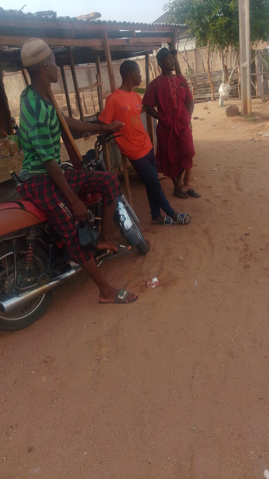 Three young men are standing around a motorbike in a dusty, outdoor area. The man on the left is wearing a green and black striped t-shirt, red plaid shorts, and a light brown cap. He is sitting on the motorbike, holding a black plastic bag. The man in the middle is wearing an orange t-shirt and dark pants, and he is leaning against a wooden structure. The man on the right is wearing a red robe with gold embroidery and a black cap. He is standing with his hands in his pockets. The environment appears to be a street or roadside, with a wooden shelter in the background and sandy ground. The overall impression is a casual, everyday scene in what appears to be a somewhat rural or less developed urban setting in Tempure, Nigeria.