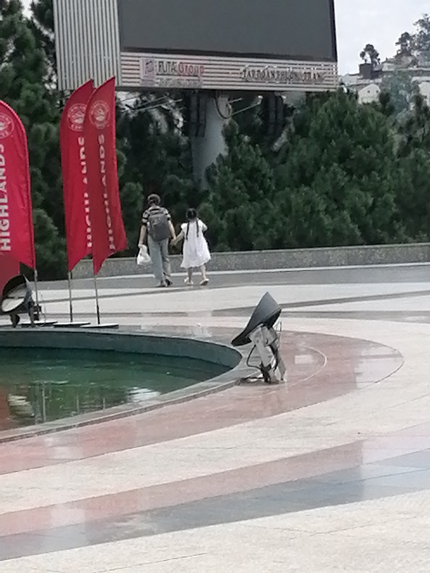 The image captures a scene in Da Lat, Vietnam, focusing on a paved area with a curved water feature in the foreground. Two red banners with white text and circular emblems are prominently displayed on the left side of the frame, flanking a person walking away from the camera. This individual is carrying a backpack and holding a white plastic bag. Beside this person, a young girl in a white dress is walking, holding the individual's hand. In the background, a large, dark billboard or screen is visible, with horizontal stripes and text. Green trees and a building structure are also discernible behind the people and banners. Further to the right, a dark conical object is positioned near the water feature, with a figure or structure partially visible beneath it. The ground is paved with light-colored tiles, some of which appear wet, reflecting the surrounding environment.