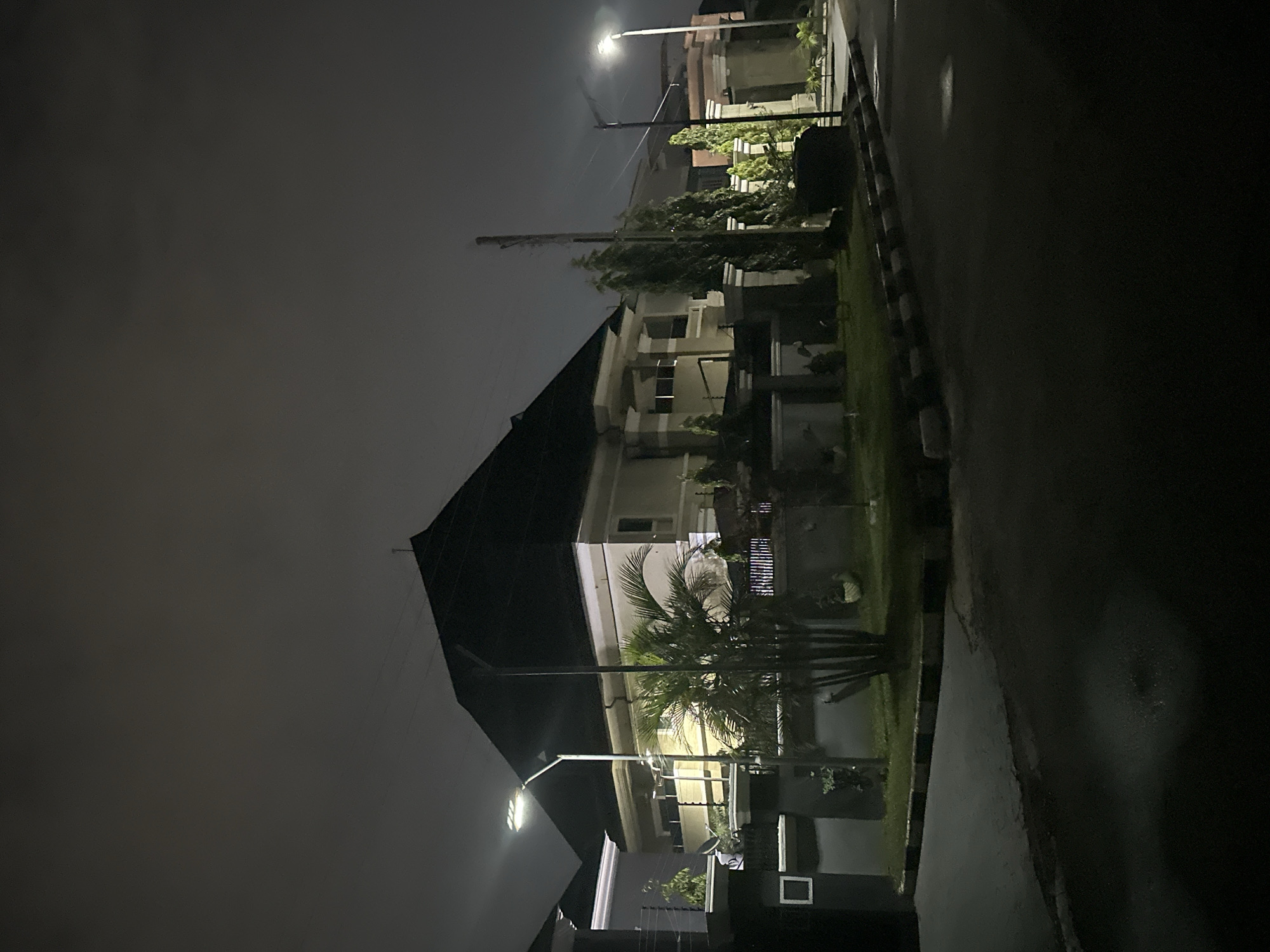 This image shows a residential street at night in Abuja, Nigeria, with a prominent two-story house featuring a dark, pitched roof. The house has cream-colored walls, balconies, and is surrounded by a low wall and fence. A street lamp illuminates the scene, casting light on a palm tree and other manicured vegetation in the foreground. The road appears wet, suggesting recent rainfall. The sky is overcast and dark, typical of nighttime conditions in the city. There are no people visible in the image.