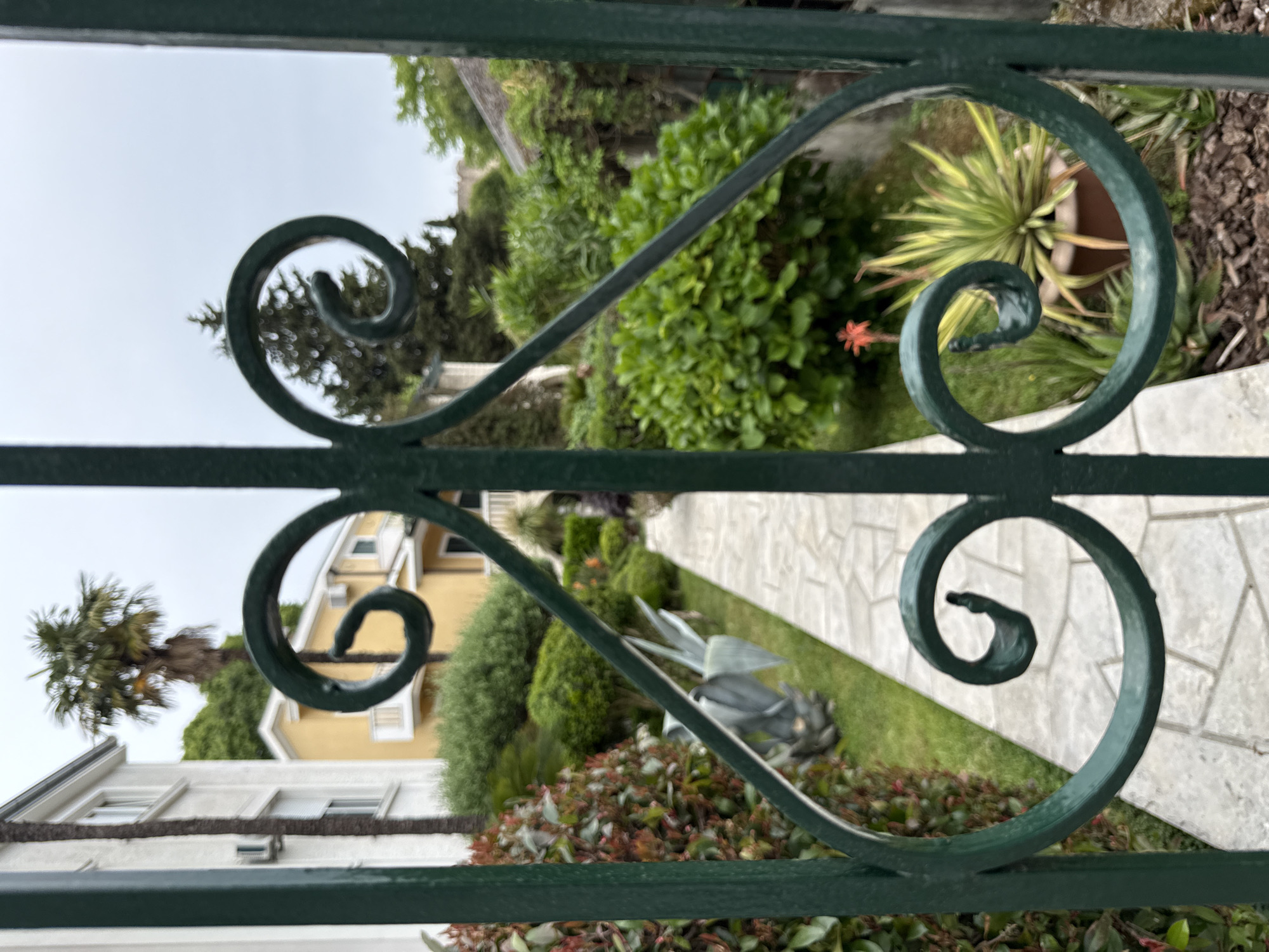 The image presents a view through a green wrought iron gate, likely located in Herceg Novi, Montenegro, based on architectural styles and landscaping. The foreground shows the ornate, curving designs of the gate. Beyond the gate, a stone-paved walkway leads towards a yellow house. The surrounding landscape includes diverse greenery: neatly trimmed bushes, flowering plants, and a palm tree visible on the left. A statue is also visible in the yard. The sky is overcast, suggesting a cloudy day.