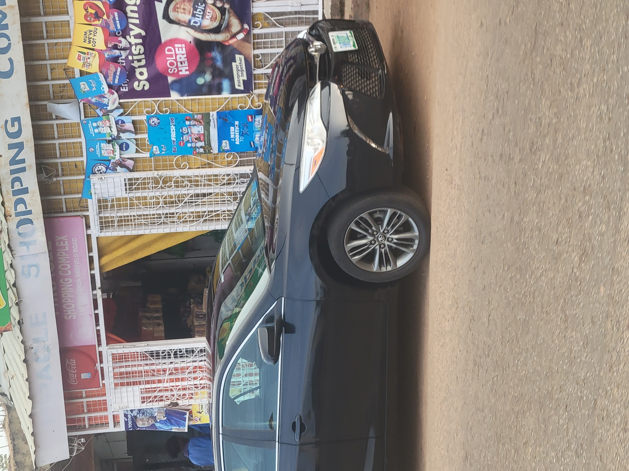 A black car is parked on a paved surface beside a retail space. The retail space has a white facade and a sign above the entrance that reads "ACE SHOPPING COMPLEX." Other signs are displayed, including ones for Coca-Cola, Nivea, and Dubic Malt.