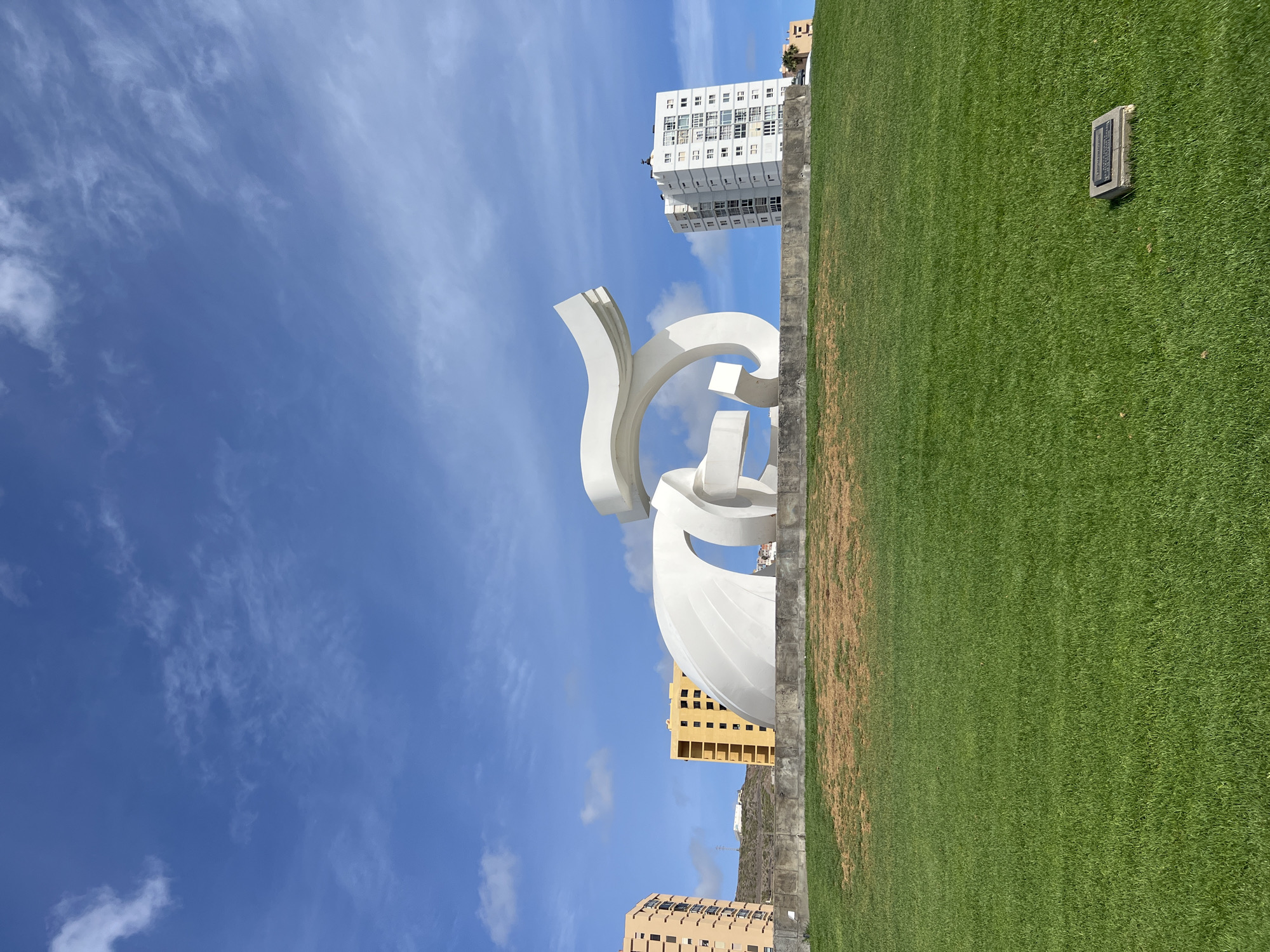 The image shows the "Monumento al Atlante" sculpture in Las Palmas, Spain, set against a blue sky with scattered clouds. The sculpture is a large, white abstract structure. Behind the sculpture are several buildings, including a tall white apartment building and other structures of varying heights. To the right of the sculpture is a green grassy area separated by a stone retaining wall. A plaque or marker is visible within the grassy area.