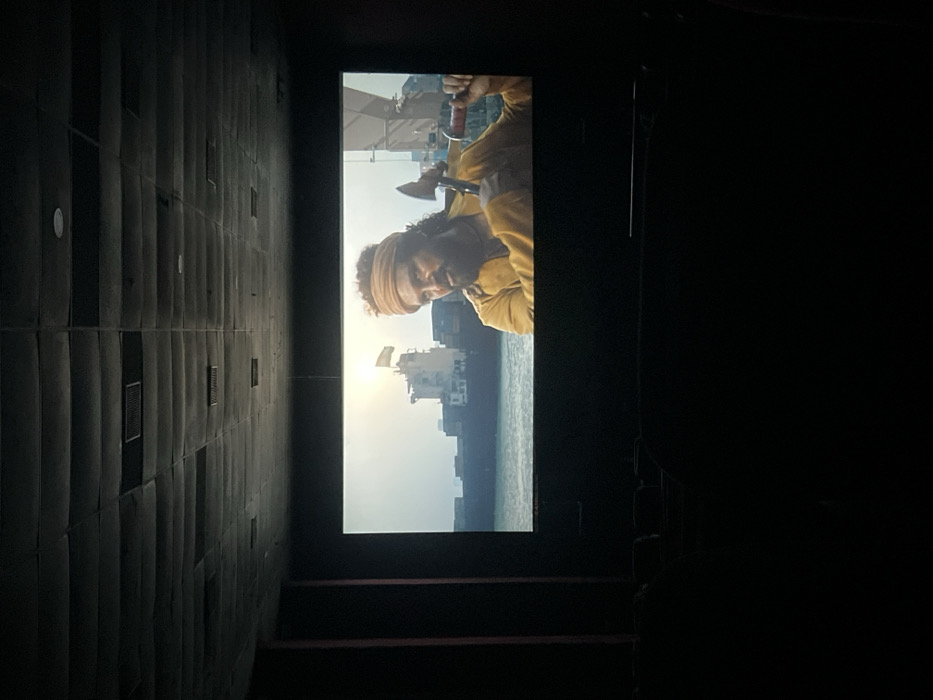 The image displays a dark movie theater, observed from a side angle, with a large film screen prominently featured. The room is dimly lit, emphasizing the brightness of the screen. The left side of the theater interior shows a dark wall with vertical, padded panels, likely acoustic, punctuated by several small rectangular vents. To the right, the cinema space is shrouded in darkness, with faint outlines of steps leading down and the tops of seating rows barely visible.
The movie screen projects a scene featuring a man with dark, curly hair, a beard, and a mustache, wearing a light-colored turban or headwrap and a yellow or mustard-colored long-sleeved garment. He appears focused, with an axe visible near his head. The background of the movie scene depicts a large cargo ship or tanker on what appears to be open water. The sun is low in the sky, creating a bright glare on the left side of the screen, suggesting either sunrise or sunset. Faint structures, possibly buildings or parts of a port city, can be seen in the distant background of the film. White structures, perhaps sails or rigging, are also visible at the top of the movie frame. The image captures the experience of watching a film in a cinema hall, possibly in Tinsukia, India. No visible text is present in the image.