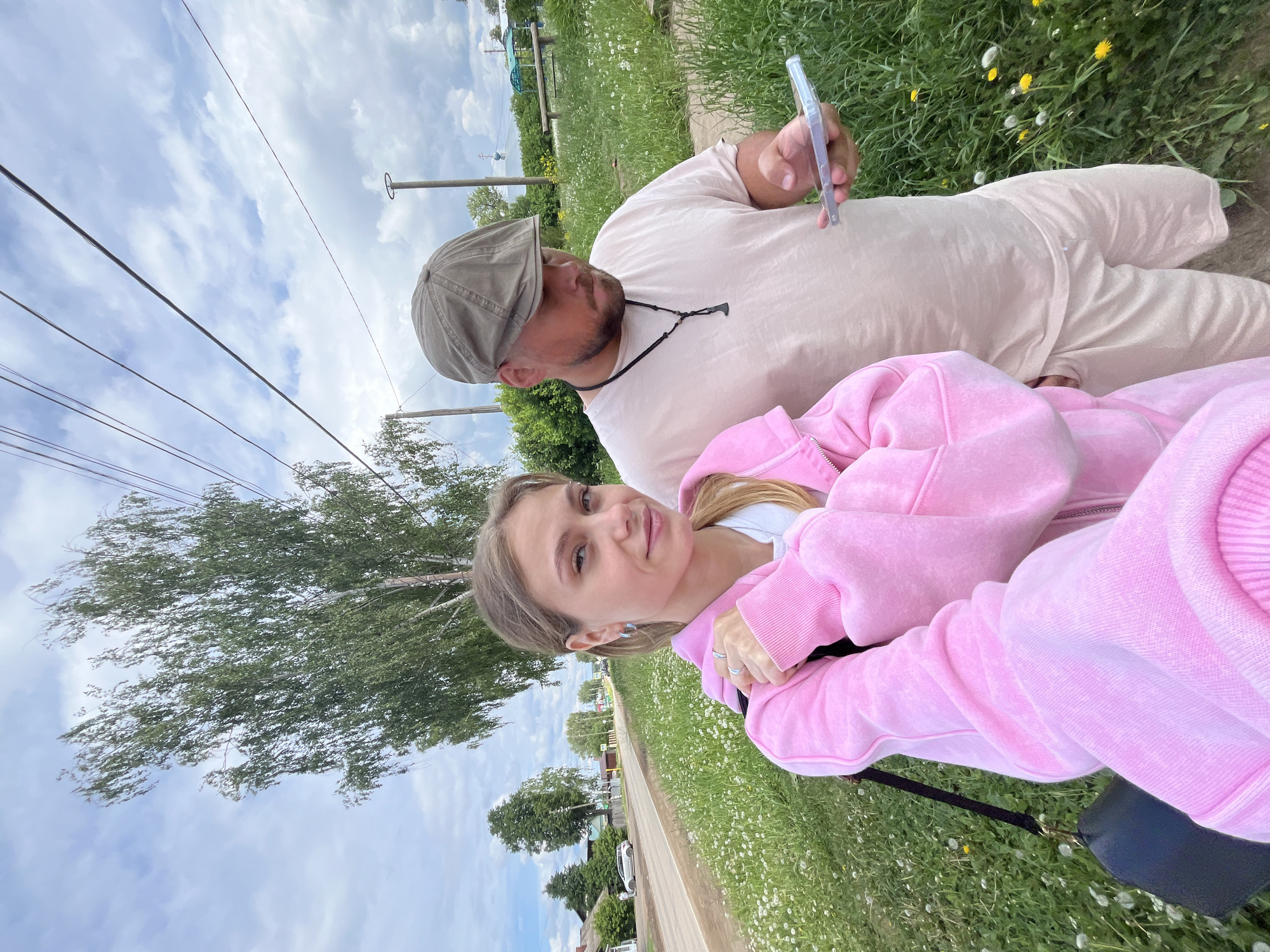 Two people are positioned outdoors on a grassy area next to a paved path. A woman with light hair is closer to the foreground, wearing a pink hooded jacket. Behind her is a man wearing a light-colored outfit and a cap, holding a smartphone. The background includes a tree, utility poles with wires, and a view of the city of Фоки, Russia. Dandelions are scattered in the grass. A road with a parked car is visible.