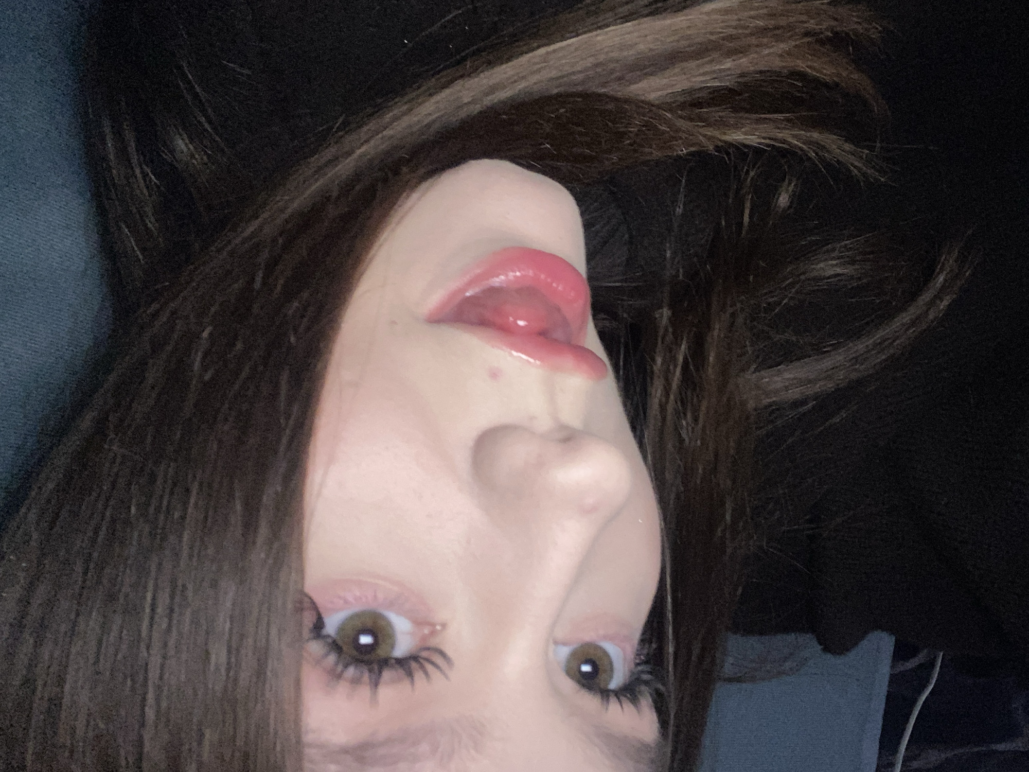 The image presents an inverted, close-up portrait of a person, likely a young woman, whose head is tilted back, revealing her face from a low angle. She looks directly into the camera with wide, warm brown eyes framed by long, dark eyelashes. Her lips are covered in shiny pink lip gloss, and her tongue is subtly extended, touching her upper lip, suggesting a playful or candid moment. A small blemish is visible below her lower lip.
Her long, dark brown hair frames her face, with strands falling across the top and sides of the frame. The background is predominantly dark, suggesting an indoor setting with subdued lighting, possibly at night. A textured blue fabric, potentially clothing or furniture, is visible on the left side of the frame. The lighting creates a slightly reflective sheen on her lips and eyes. There are no discernible objects beyond the person and minimal background elements. No text is visible in the image. The image itself offers no specific visual cues to indicate the location context of Chaikovsky, Russia.