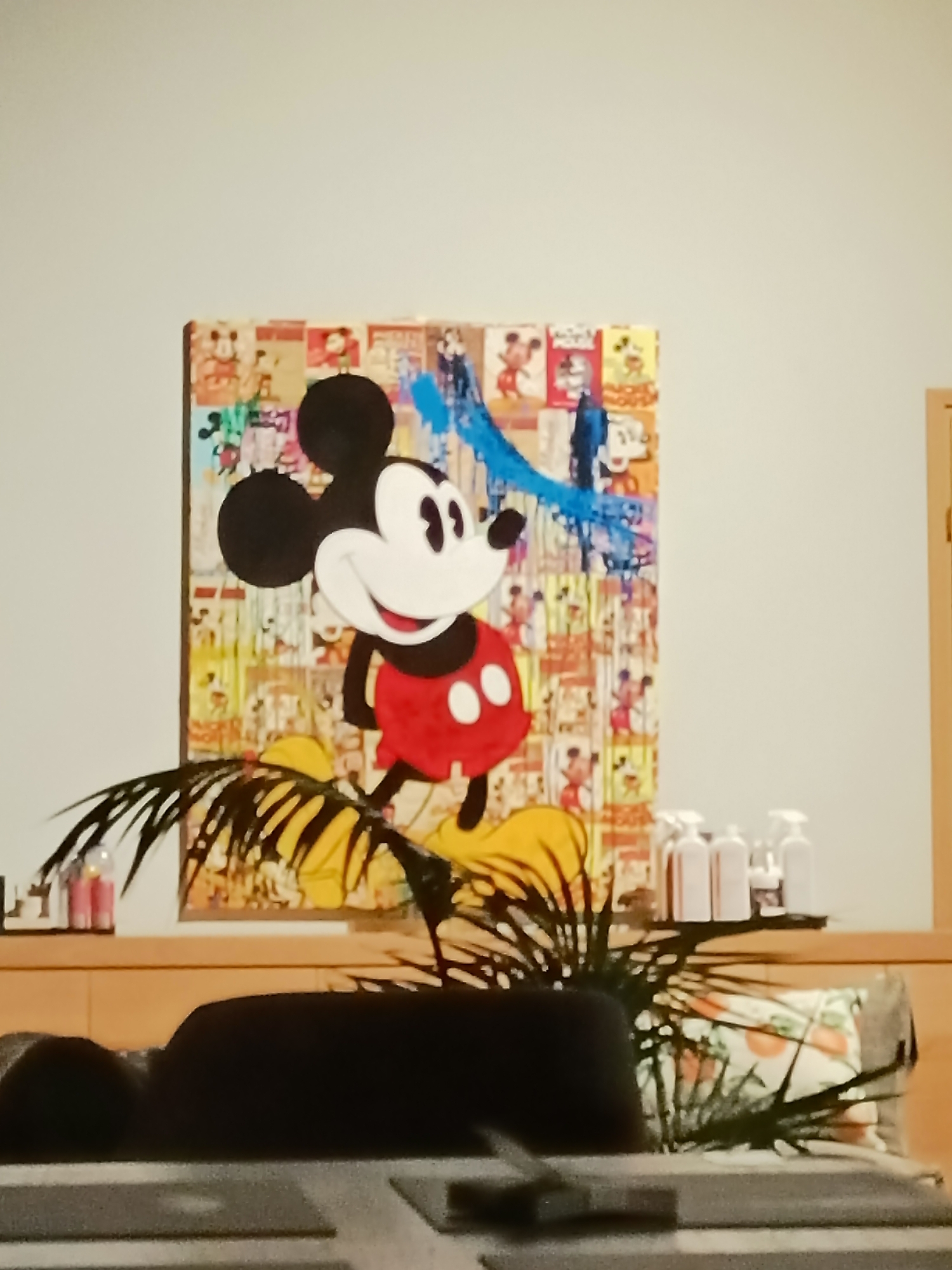 The image shows a painting of Mickey Mouse on a wall, with a plant in the foreground. The painting is a collage of Mickey Mouse images in various poses and colors, with a large, cartoonish depiction of Mickey Mouse in the center. The background of the painting appears to be made up of older comic book pages or posters featuring Mickey Mouse. A bright blue abstract shape is painted over parts of the collage. The Mickey Mouse figure in the center has a large head with prominent ears, a wide smile, and is wearing a red vest and yellow shorts.
In front of the painting, a dark green, leafy plant is positioned. The plant's fronds partially obscure the lower portion of the painting and extend towards the viewer. Behind the plant, on a wooden surface, are several bottles, likely cleaning supplies, including spray bottles. To the right of the bottles, a portion of a wall and a door frame are visible.
The overall scene appears to be indoors, possibly in a home or office. There are no people clearly visible in the image, though a dark, blurred object in the lower foreground might be furniture or a person's clothing.
The location of the image in Tallinn, Estonia, cannot be determined from the visual information provided in the image itself.