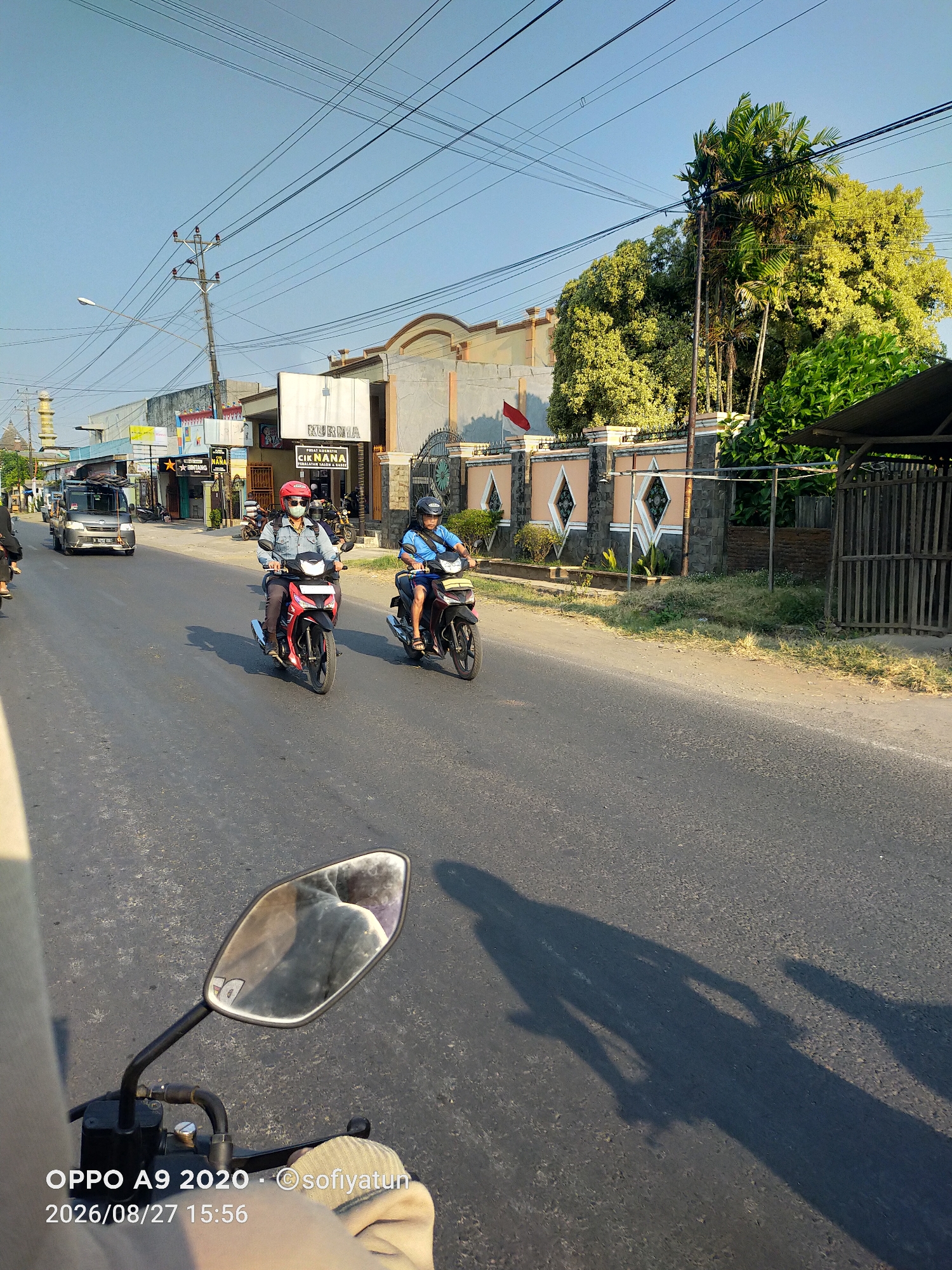 The image shows a street scene in Tayu, Indonesia, on a sunny day. Two individuals are riding motorcycles. The person in the foreground on the left is wearing a red helmet and a denim jacket, riding a red motorcycle. The person on the right is wearing a blue shirt and a black helmet, riding a dark-colored motorcycle. Both riders are wearing face masks. Several buildings line the street, including one with signage that reads "KURMA" and "CIK NANA PERALATAN SALON & SPA". A red and white Indonesian flag is visible on a pole near one of the buildings. Power lines are strung across the sky. The road is asphalt, and shadows of the motorcycles and riders are cast on the surface. The lower right corner of the image shows a reflection in a motorcycle's side mirror, revealing the back of the photographer's head and shoulder, and part of their clothing. The timestamp on the image indicates it was taken on August 27, 2026, at 15:56.