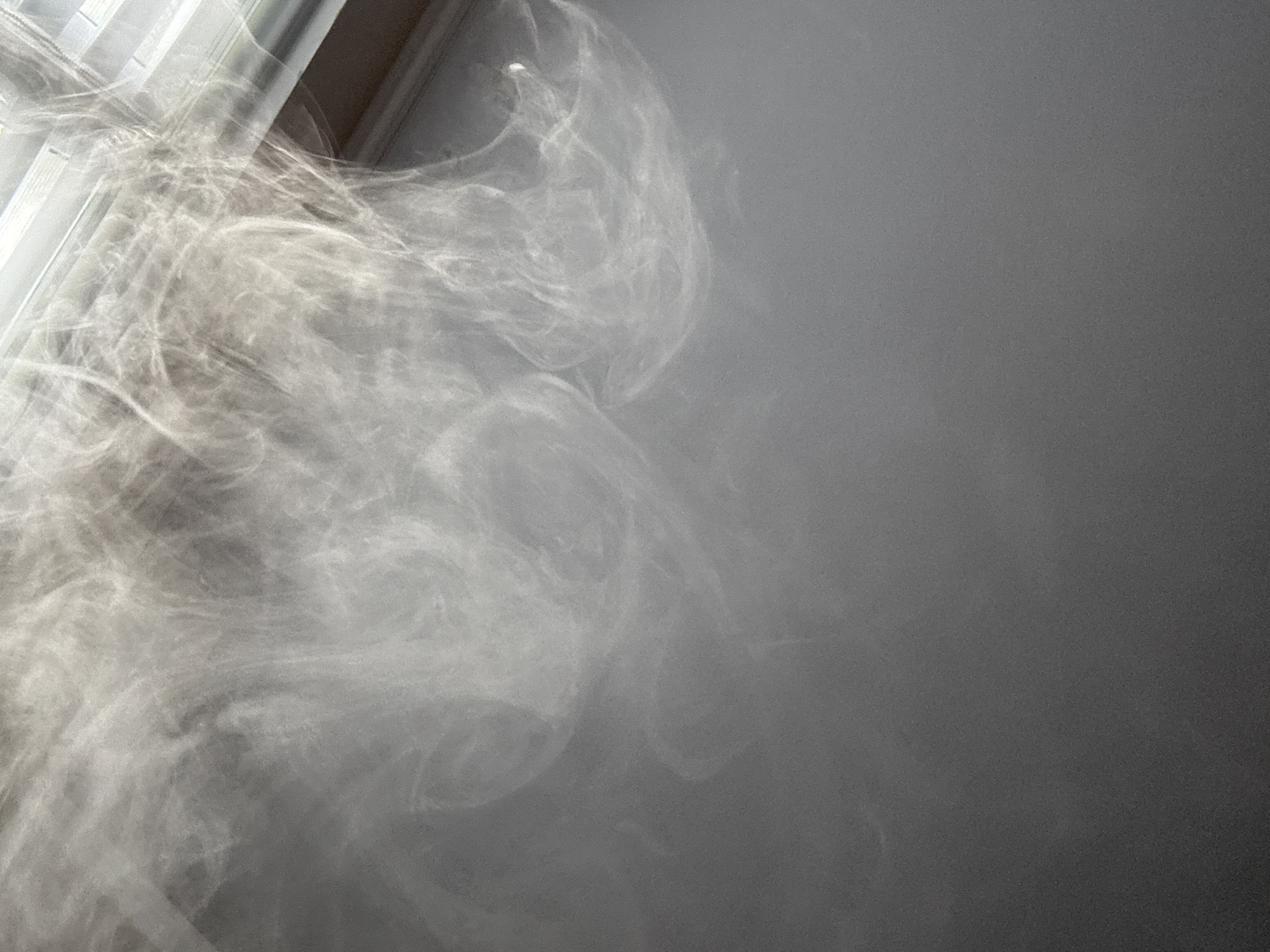 The image shows a close-up view of smoke or steam, illuminated by light filtering through what appears to be blinds. The smoke is wispy and ethereal, forming swirling patterns against a plain gray background. The light creates bright highlights and soft shadows, adding depth and texture to the composition. There are no visible people or objects other than the smoke and the implied environment of a room with a window. No specific location in Saint Petersburg, Russia can be identified from this image.