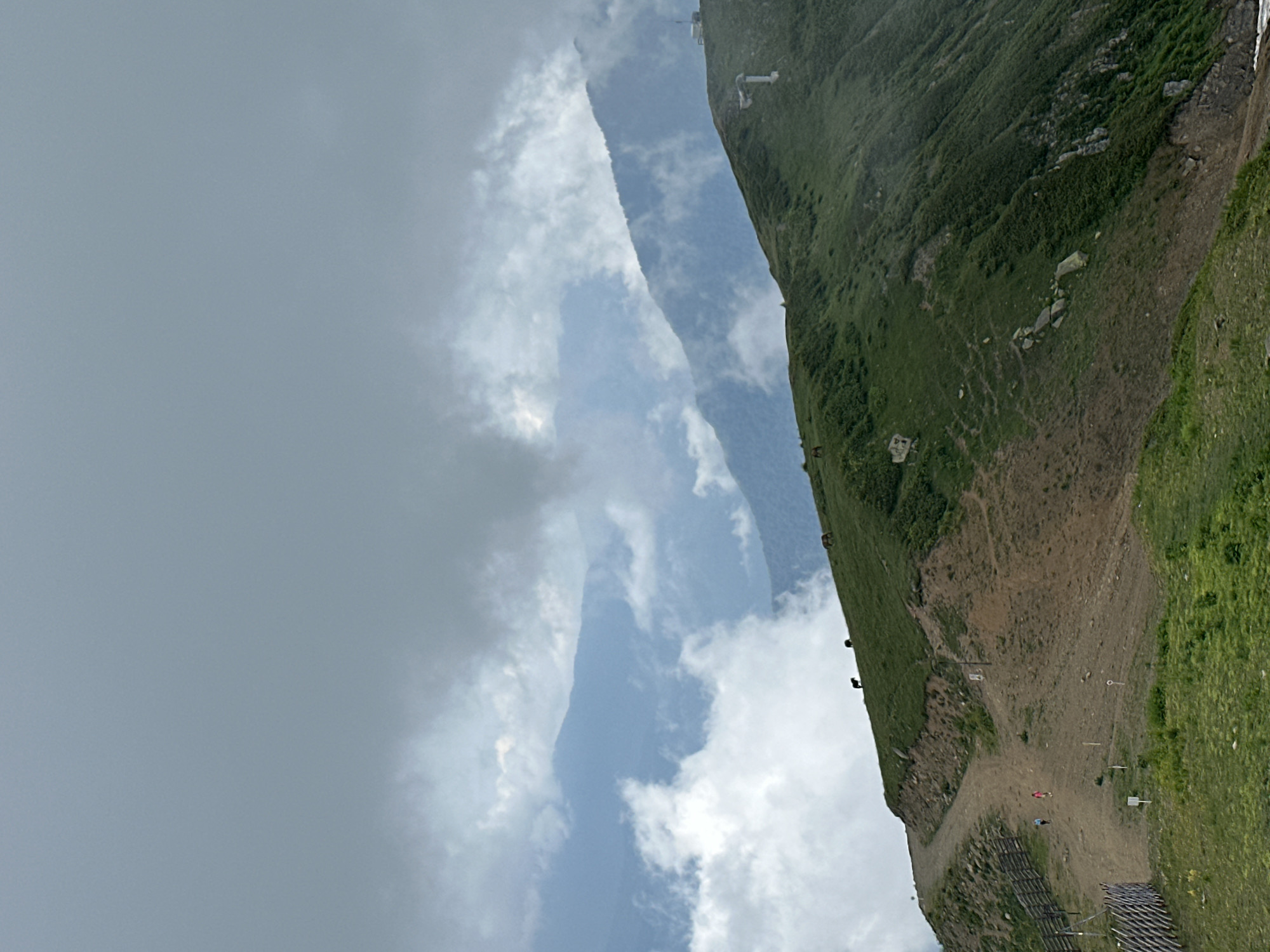 The image depicts a mountain landscape in Estosadok, Russia, under a cloudy sky. In the foreground, a dirt path winds up a steep, green, grassy slope. Two people, one wearing a pink shirt and the other a blue shirt, are walking up the path. A wooden fence runs along the left side of the path. Further up the slope, several dark-colored animals, possibly horses, are grazing on the grass. In the background, layers of mountains are visible, partially obscured by clouds and mist, creating a sense of depth and atmospheric perspective. On the crest of one of the further mountains, a structure, possibly a telecommunication tower or a building, is discernible. The overall scene conveys a rugged, natural environment with signs of human presence, such as the path and the people.