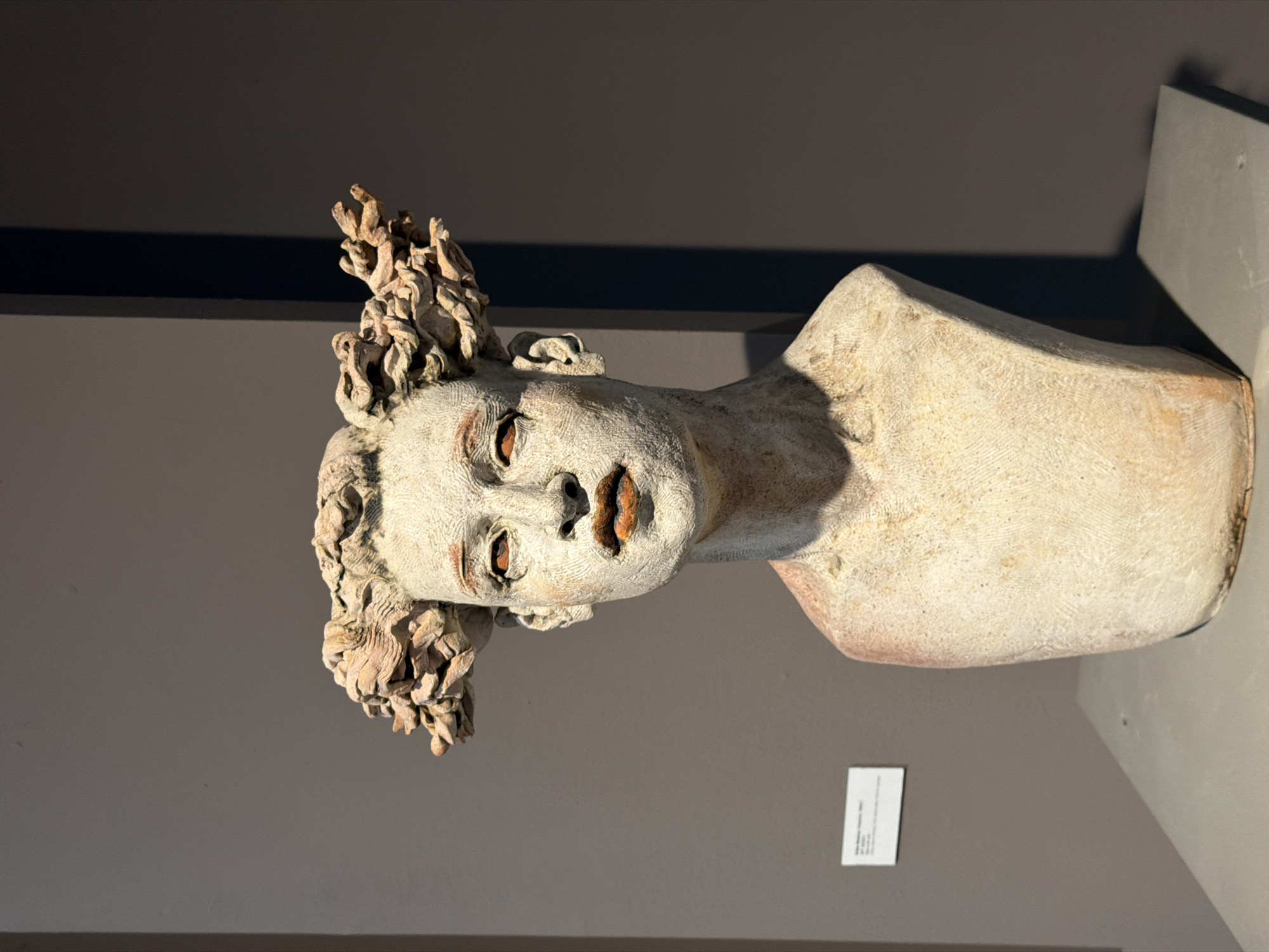 A bust of a woman with textured, light-colored skin and sculpted, wavy hair is displayed on a gray surface against a dark gray wall. The sculpture features defined facial features with an orange hue on the lips and around the eyes. A small white label is affixed to the wall to the left of the sculpture. The lighting casts a shadow to the right of the bust. This artwork is located in the Picasso Museum in Barcelona, Spain.