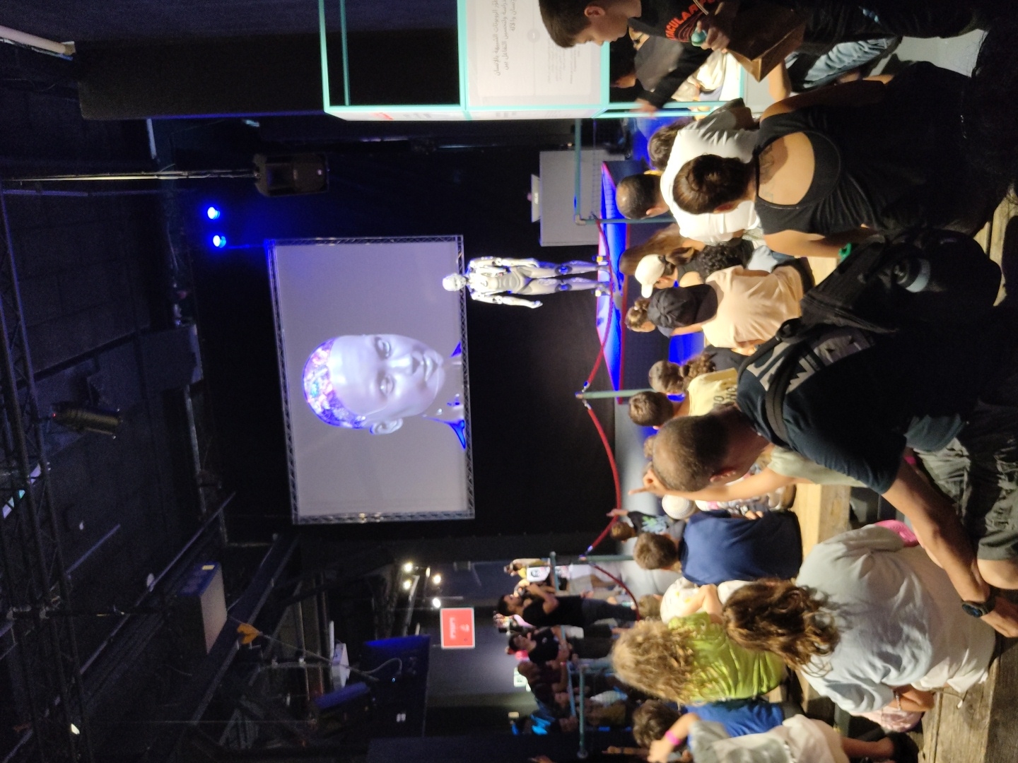 The image captures an indoor event likely at the Technion in Haifa, Israel, featuring a presentation or demonstration involving robotics and AI. In the background, a large screen displays a realistic, digitized head of a person with intricate patterns on their scalp. In front of the screen, a life-sized humanoid robot stands on a raised platform, illuminated by blue lights.
The foreground is occupied by an audience seated on tiered wooden platforms, facing the presentation. The audience consists of a diverse group of people, including children and adults, seated closely together. Some individuals are actively engaged, with one child pointing towards the robot. The environment appears to be a dark hall, possibly a lecture hall or auditorium, with visible lighting fixtures and structural elements in the ceiling. A red rope barrier separates the audience from the presentation area. To the right, a display board with Arabic text is visible, suggesting a multilingual context for the event. The overall atmosphere suggests an educational or exhibition setting focused on technological advancements.