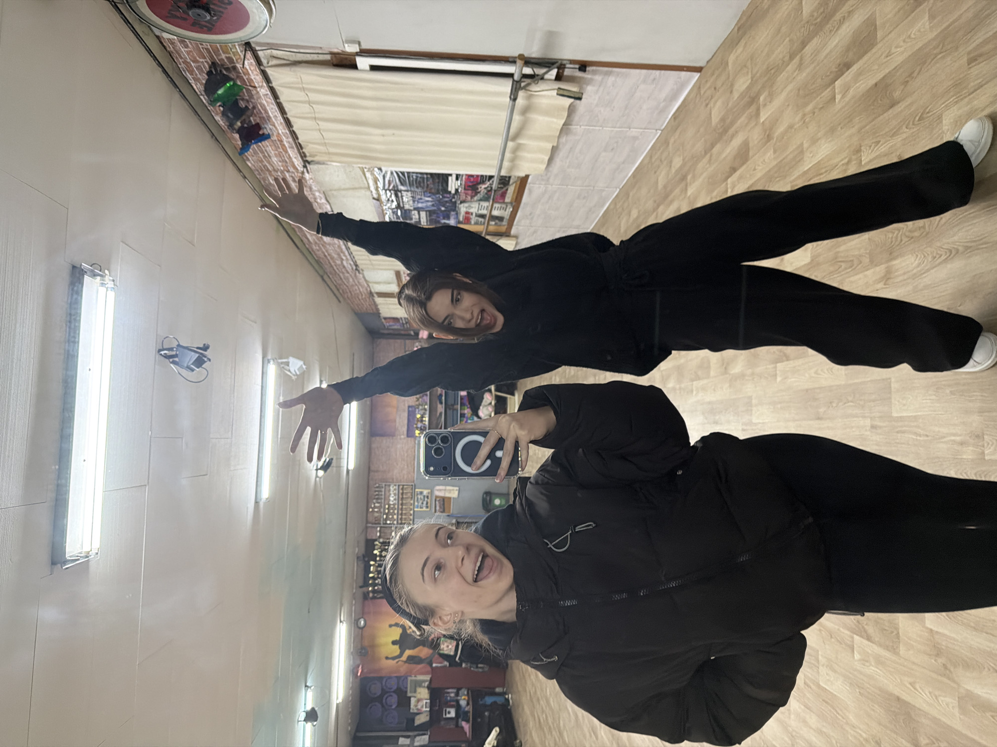 The image is a reflection of two young women taking a selfie in what appears to be a dance studio or rehearsal space in Plovdiv, Bulgaria. The studio has a wall-length mirror where the reflection is seen. The woman closest to the mirror is holding a smartphone up to take the photo, wearing a dark puffer jacket and black leggings, with her hair pulled back by a headband. She is smiling with her mouth open. Behind her in the reflection is another woman in a black sweatshirt and pants, with both arms outstretched, smiling and yelling. The studio has a wood-like floor, and fluorescent lights on the ceiling. A few items (posters, speakers) are visible on the back wall in the reflection. The overall impression is of a casual moment, likely during dance practice.