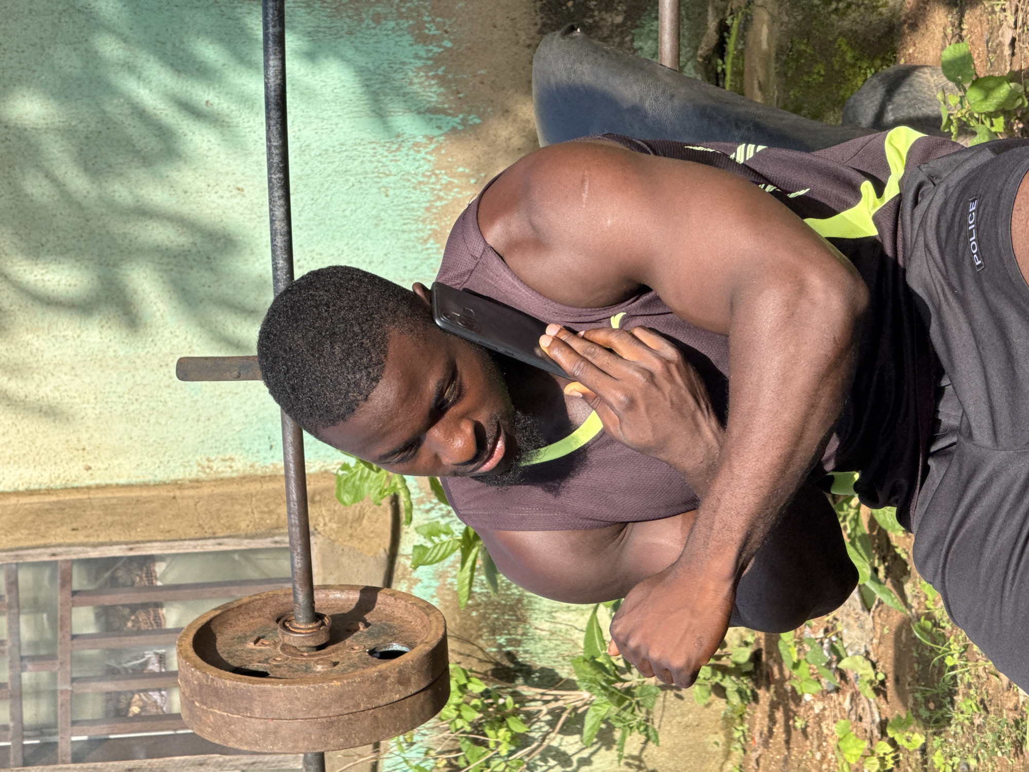 A muscular Black man, wearing a dark tank top with neon green accents and black athletic shorts with "POLICE" printed on the left thigh, is seated on a weight bench outdoors. He is holding a black smartphone to his right ear with his right hand while his left arm is bent and resting on his chest. He appears to be engaged in a phone conversation. Behind him, a rusty barbell with a weight plate is visible, positioned on a weight rack. The background consists of a weathered, light green wall with shadows cast upon it, some greenery, and a window with a metal grate. The scene is illuminated by natural sunlight, creating strong highlights and shadows. The setting suggests an outdoor workout area, potentially in a residential yard, given the presence of the house wall and surrounding vegetation. This location is identified as Akwanga, Nigeria.