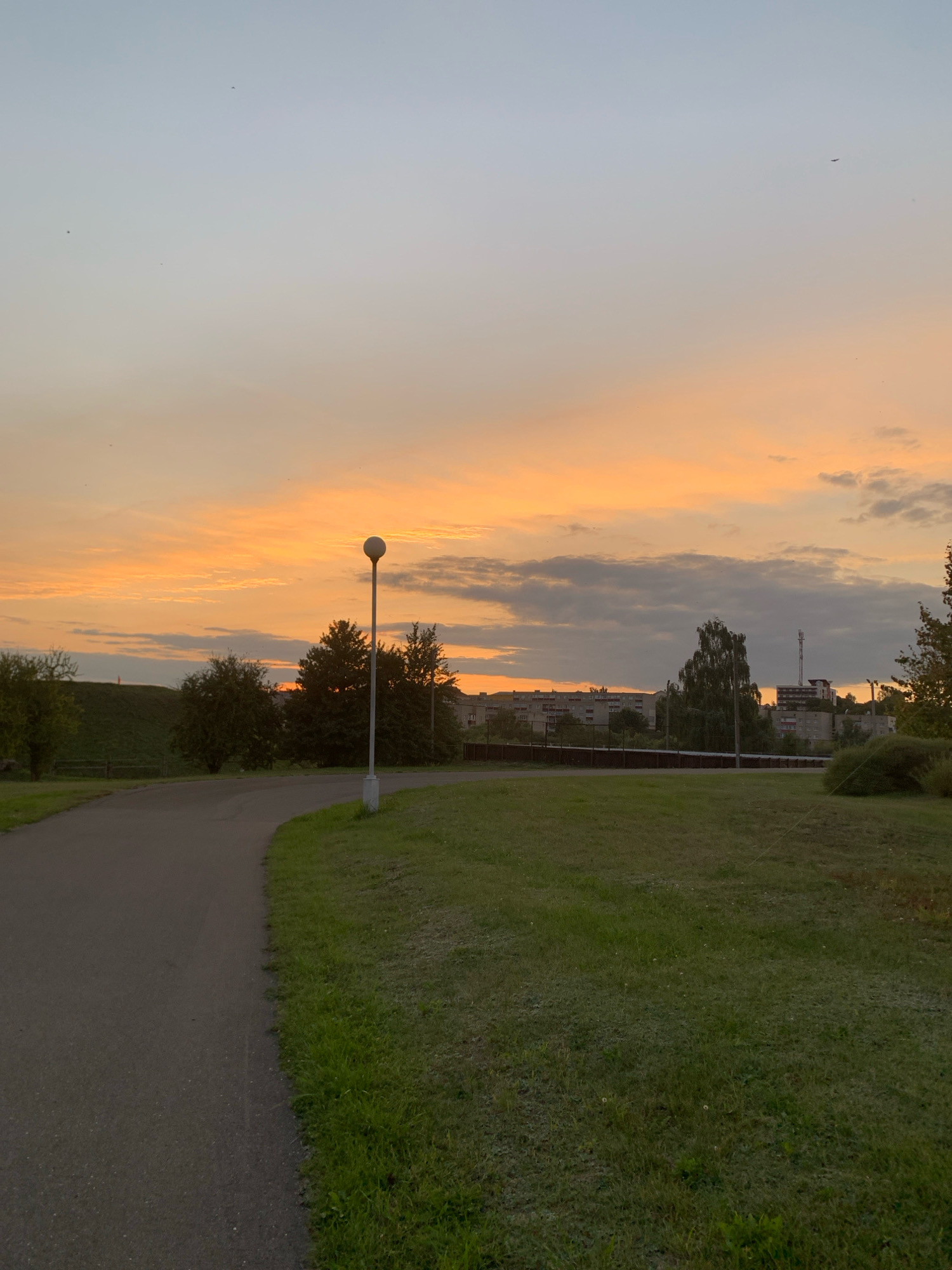 The image depicts a sunset scene in the city of Kapyĺ, Belarus. A paved pathway curves through a grassy park area, leading towards a line of trees and buildings in the background. A solitary lamppost stands beside the pathway. The sky is filled with a gradient of colors, transitioning from a pale blue at the top to a vibrant orange and yellow near the horizon, with scattered clouds. Several small birds are visible in the sky. In the distance, apartment buildings and a cell tower are present. The overall impression is one of a tranquil evening in a suburban or park setting.