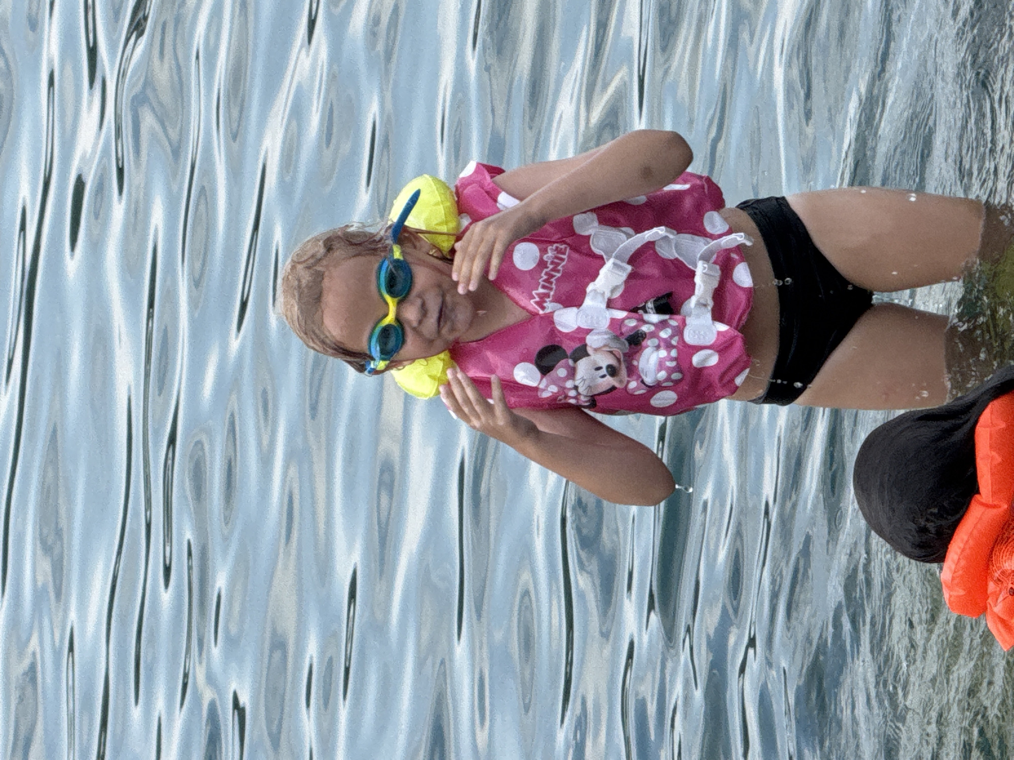 In this image, a young girl is in the water, wearing a pink and white polka dot life vest adorned with a Minnie Mouse print. She has on blue swimming goggles. Her black swimsuit bottoms are visible. Another person, whose head and shoulders are visible on the right side of the frame, is wearing an orange life vest. The water has a rippled texture, suggesting movement and light reflection. The scene appears to be outdoors, likely during the daytime, with the lighting and clear water indicating pleasant weather. The overall mood seems relaxed and playful, consistent with water activities. There is text visible on the life vest that reads "MINNIE". The location is identified as Partenit, Ukraine.