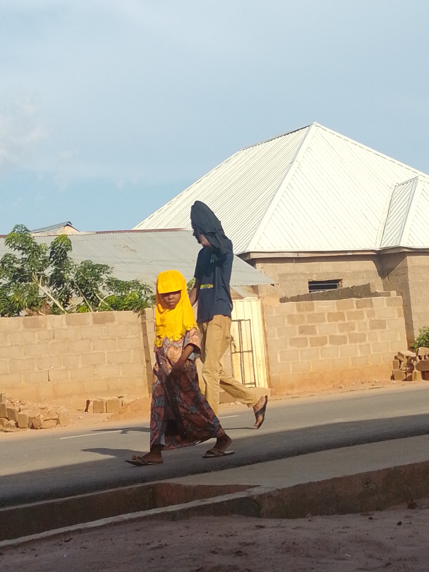 Two individuals are walking on a paved road in Tempure, Nigeria. The person in front, appearing to be a child, is wearing a bright yellow headscarf and a patterned dress. They are walking from left to right. Behind them, a taller individual, possibly an adult, is walking in the same direction, wearing a dark headscarf and tan pants. They are positioned slightly to the right of the child. The background consists of a light blue sky with some clouds, a white corrugated metal roof of a building, a brick wall, and some green foliage. The road surface is asphalt with a white line marking its edge. The lighting suggests it is daytime, with the sun casting shadows on the road.