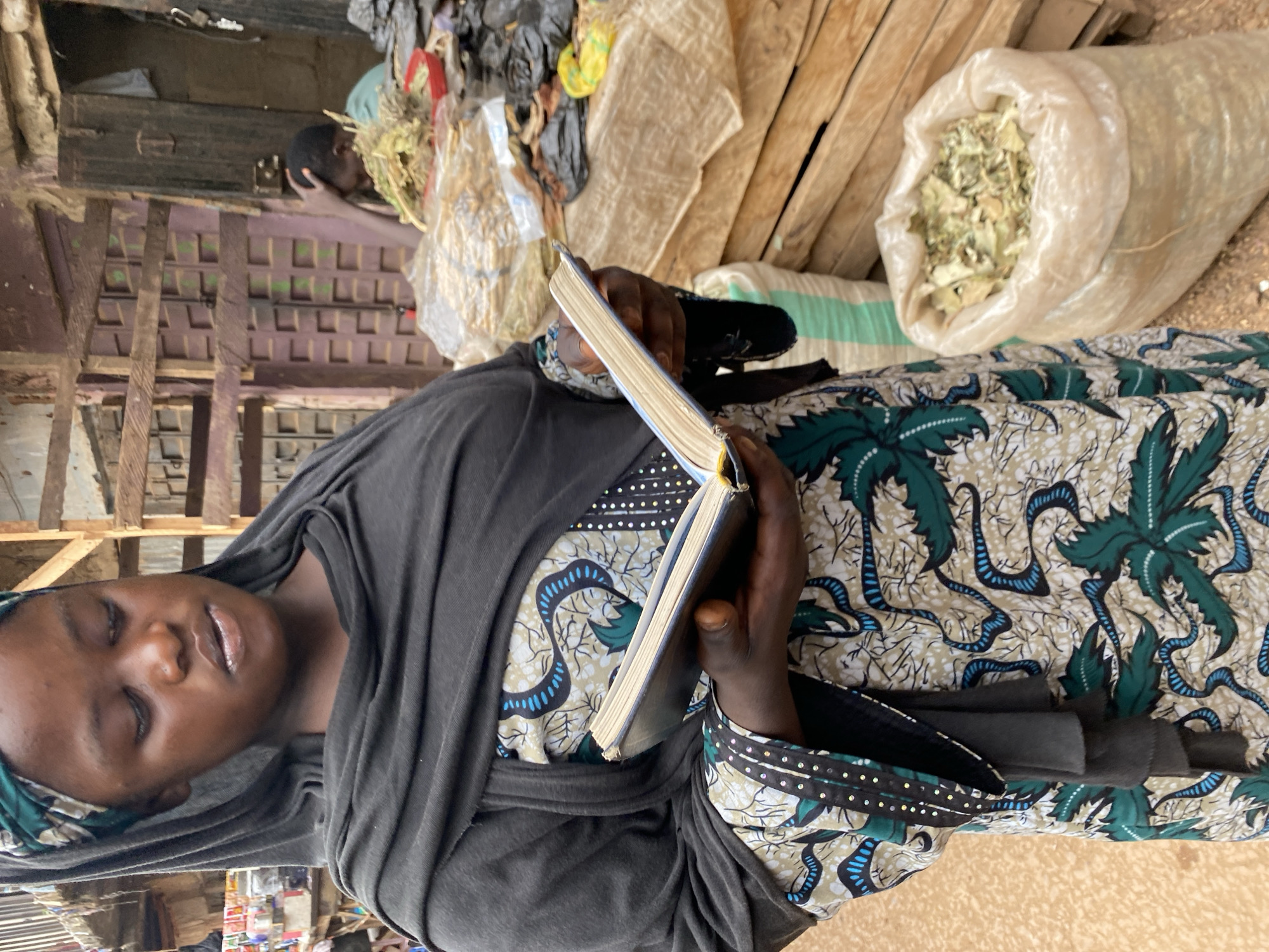 A young woman, appearing to be of Nigerian ethnicity based on the location provided, is standing in what seems to be an outdoor market in Bukuru, Nigeria. She is wearing a dark headscarf, a dark gray shawl draped over her shoulders and chest, and a colorful dress with a pattern of green leaves and blue, snake-like designs. In her hands, she is holding an open book, with the pages turned towards the viewer, and her gaze is directed downwards, as if reading or studying it. The background suggests a bustling market environment with wooden structures, shelves, and various goods for sale, including sacks of dried herbs or other produce. Another person is partially visible in the background, stooped over. The ground appears to be dirt.