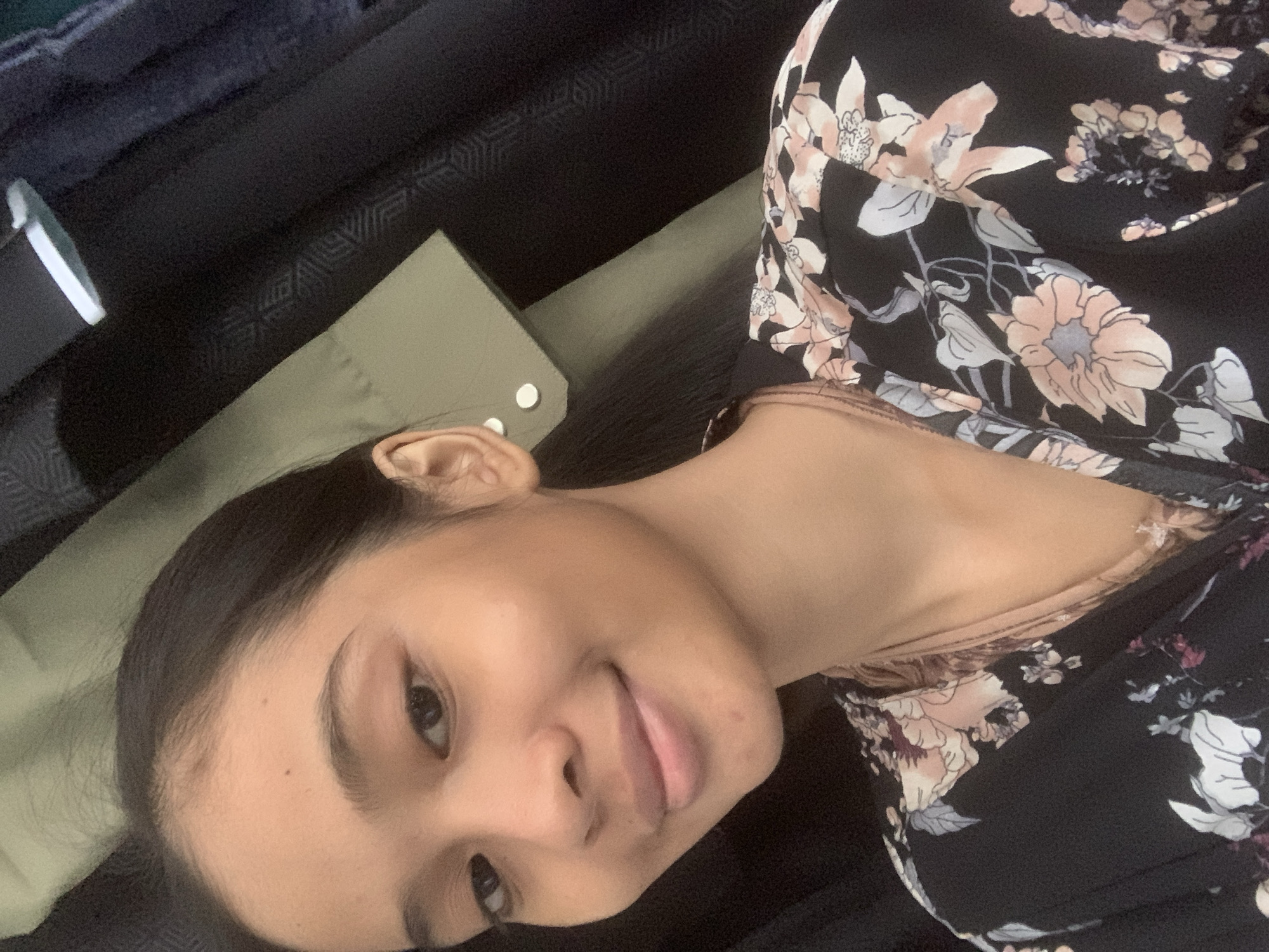 A young woman with dark hair pulled back is shown in a close-up, selfie-style portrait. She is smiling slightly and looking towards the camera. She is wearing a black, floral-patterned top with light pink and white flowers. Behind her, there are dark fabrics, possibly clothing or curtains, and a section of a light olive-colored fabric with two small white buttons. The lighting suggests an indoor setting. The image does not contain any information indicating a specific location in Sungai Meriam, Indonesia.